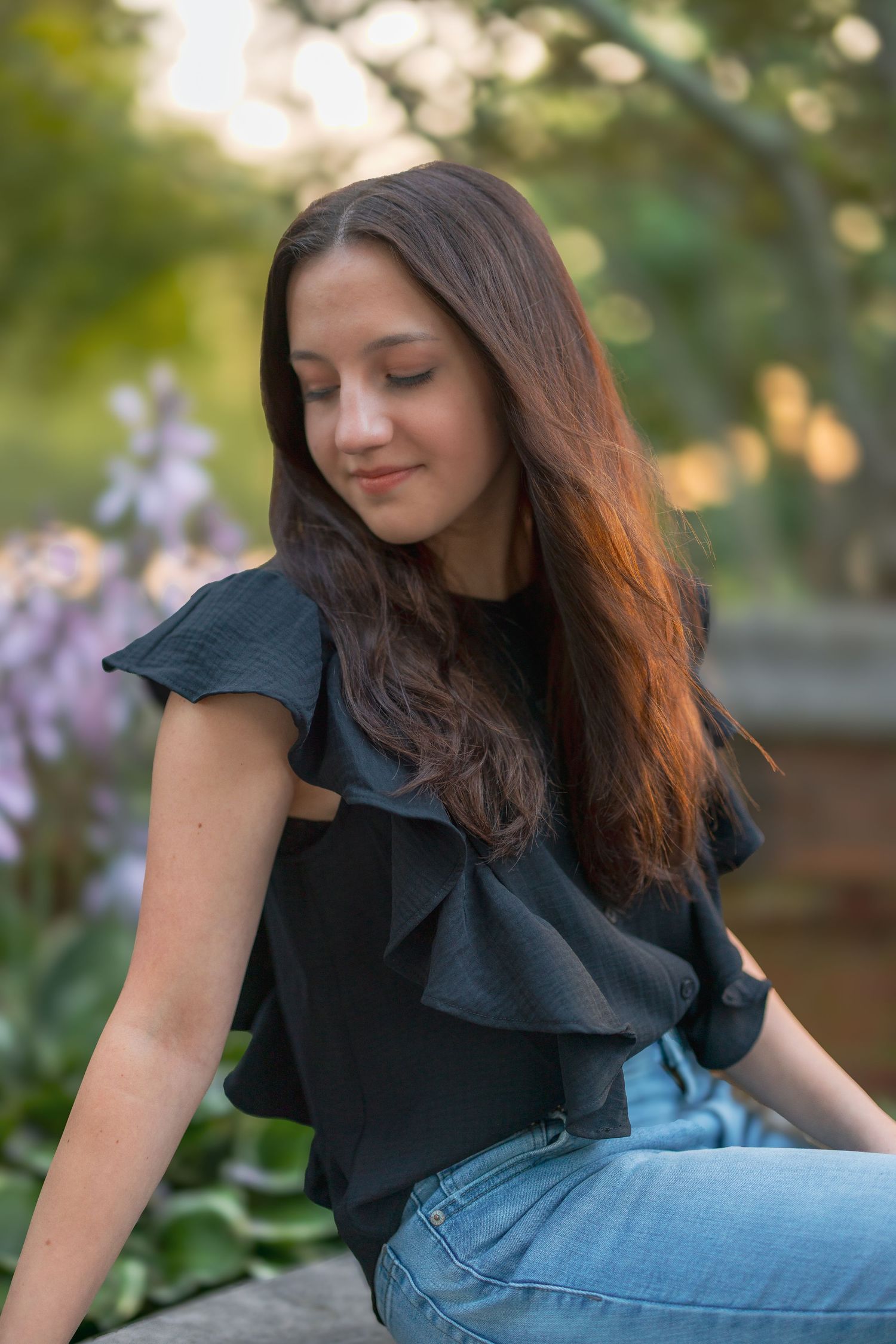 A woman in a black ruffle top and jeans poses outdoors in a series of casual portraits near blooming flowers.