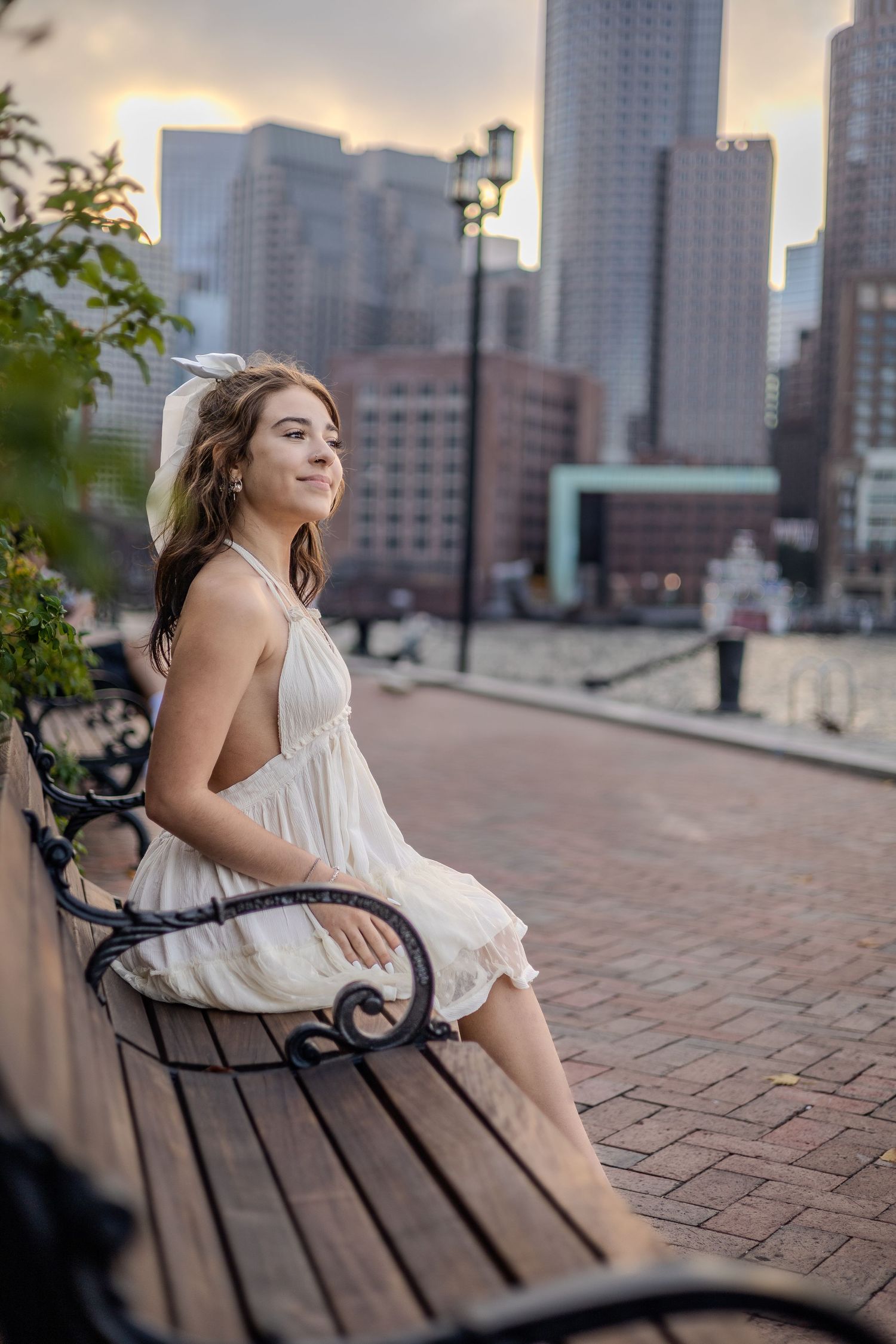 A young woman in a white sundress sits pensively on a bench against a city skyline at sunset.