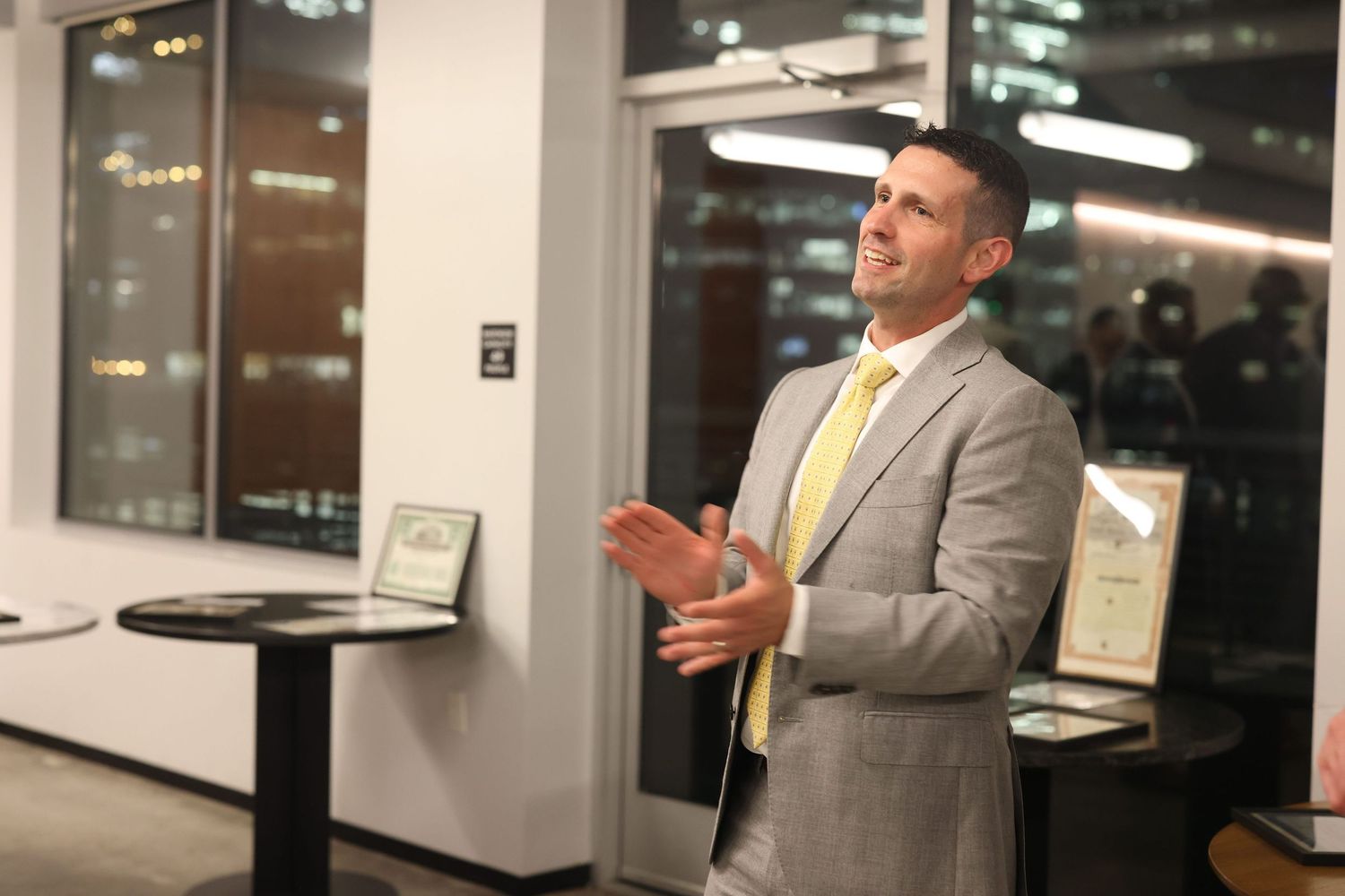 A person in a gray suit gestures while speaking in a modern office setting with glass walls.