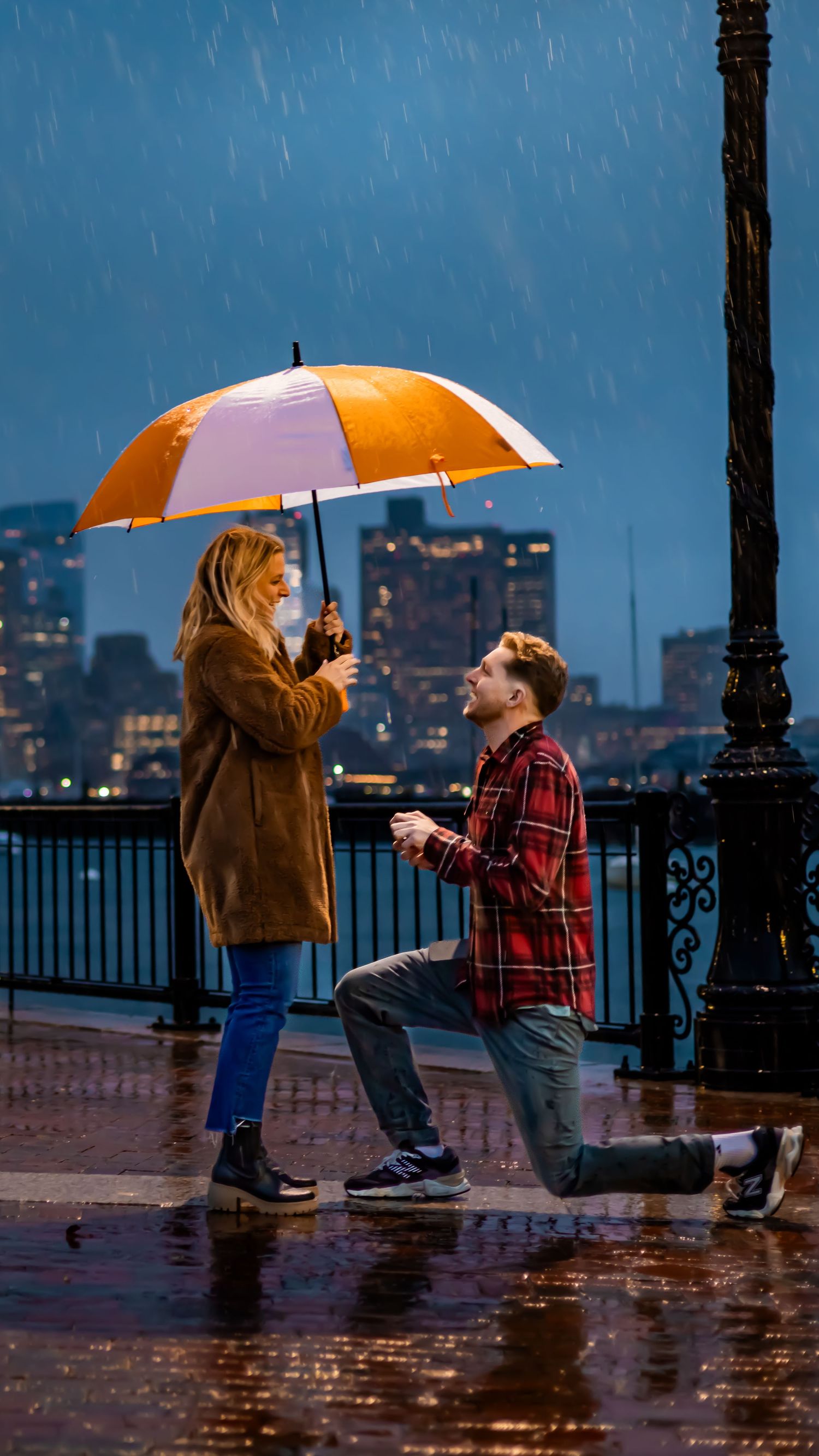 A romantic proposal scene at night in the rain with an orange umbrella and city skyline in the background.