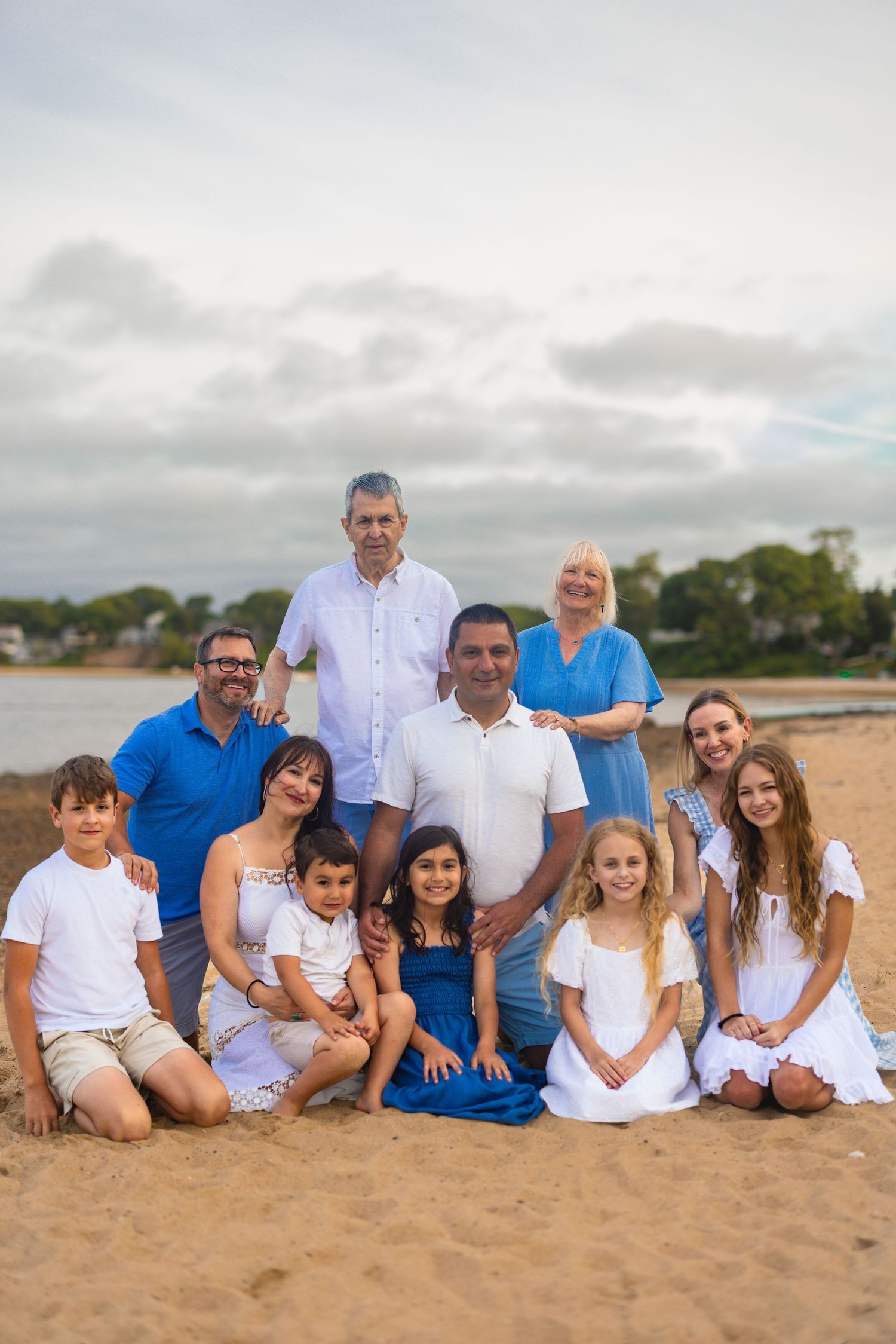 Extended family wearing blue and white outfits sits together on tropical beach sand during cloudy day.
