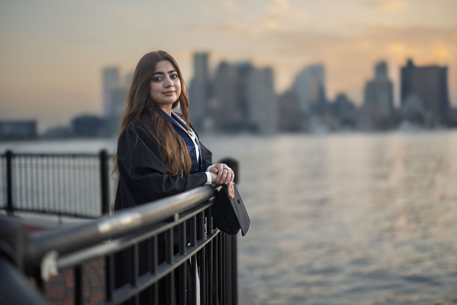 A person stands at a waterfront railing during sunset with a cityscape silhouetted in the background.