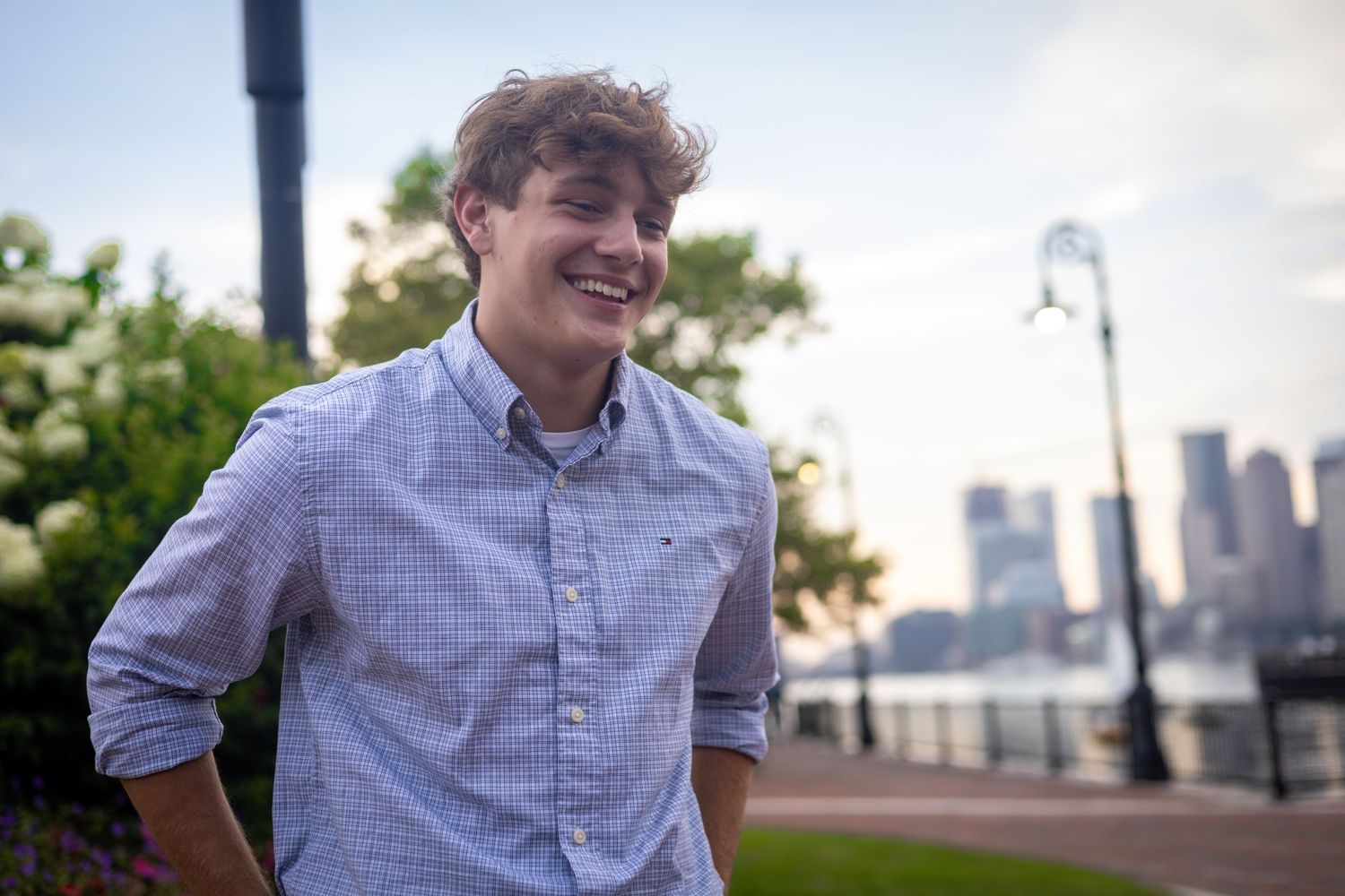Young man in checkered button-down shirt laughing and walking in an urban park with city skyline visible.