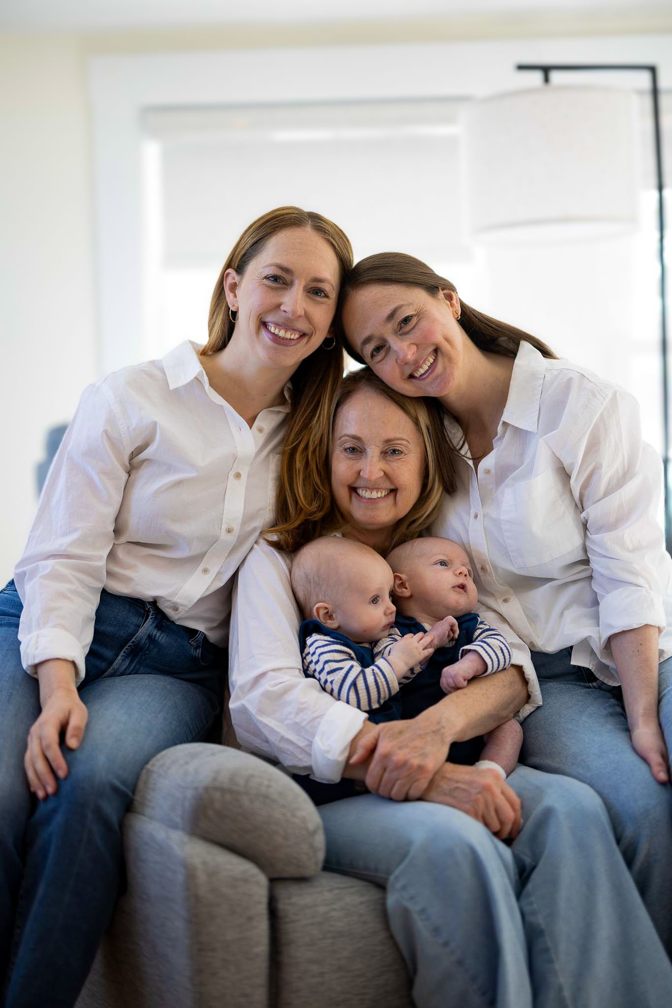 Family in matching white shirts sitting together on a grey couch, including two babies in striped outfits.