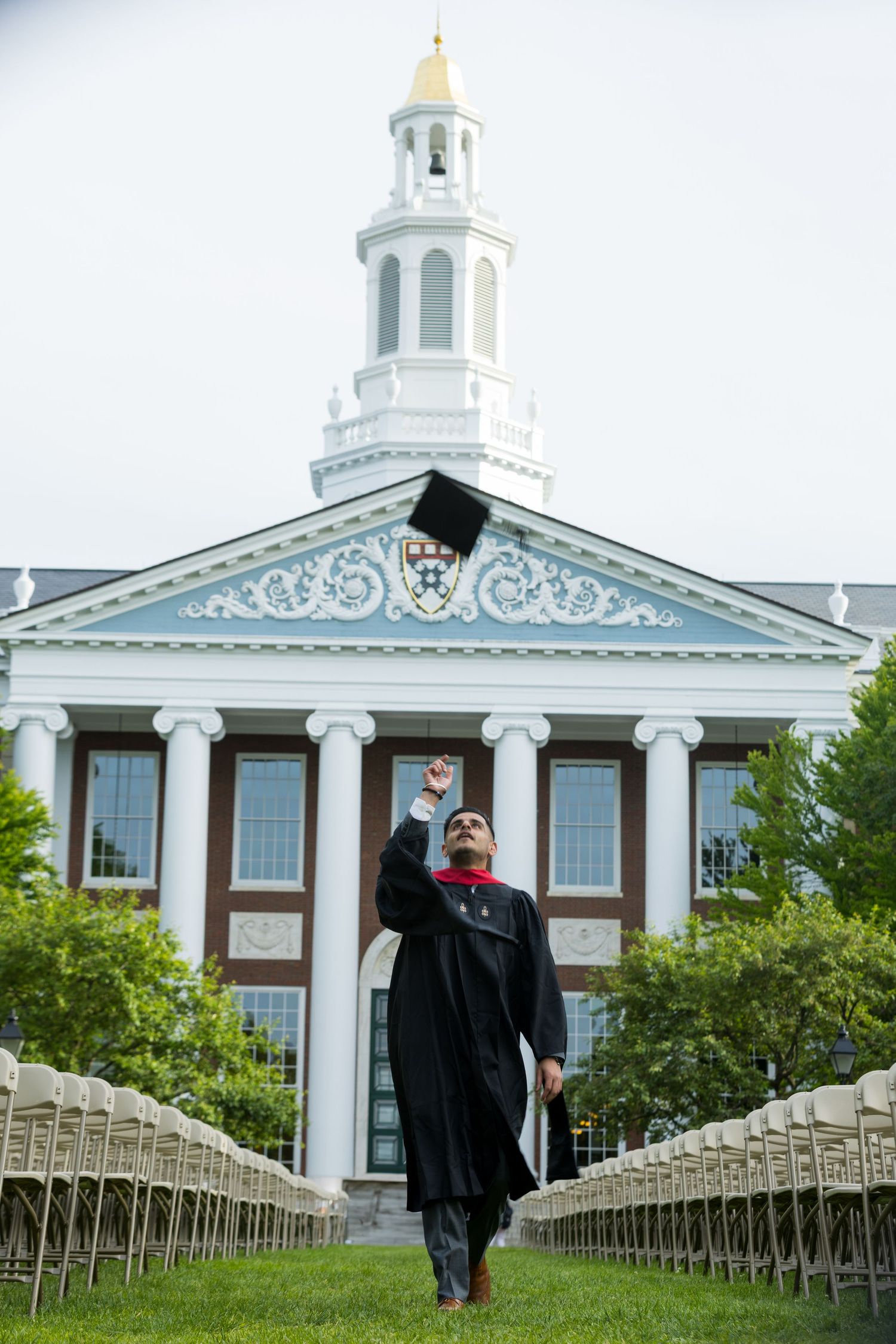 Graduate in cap and gown celebrates on steps in front of historic Harvard Business School building with white columns.