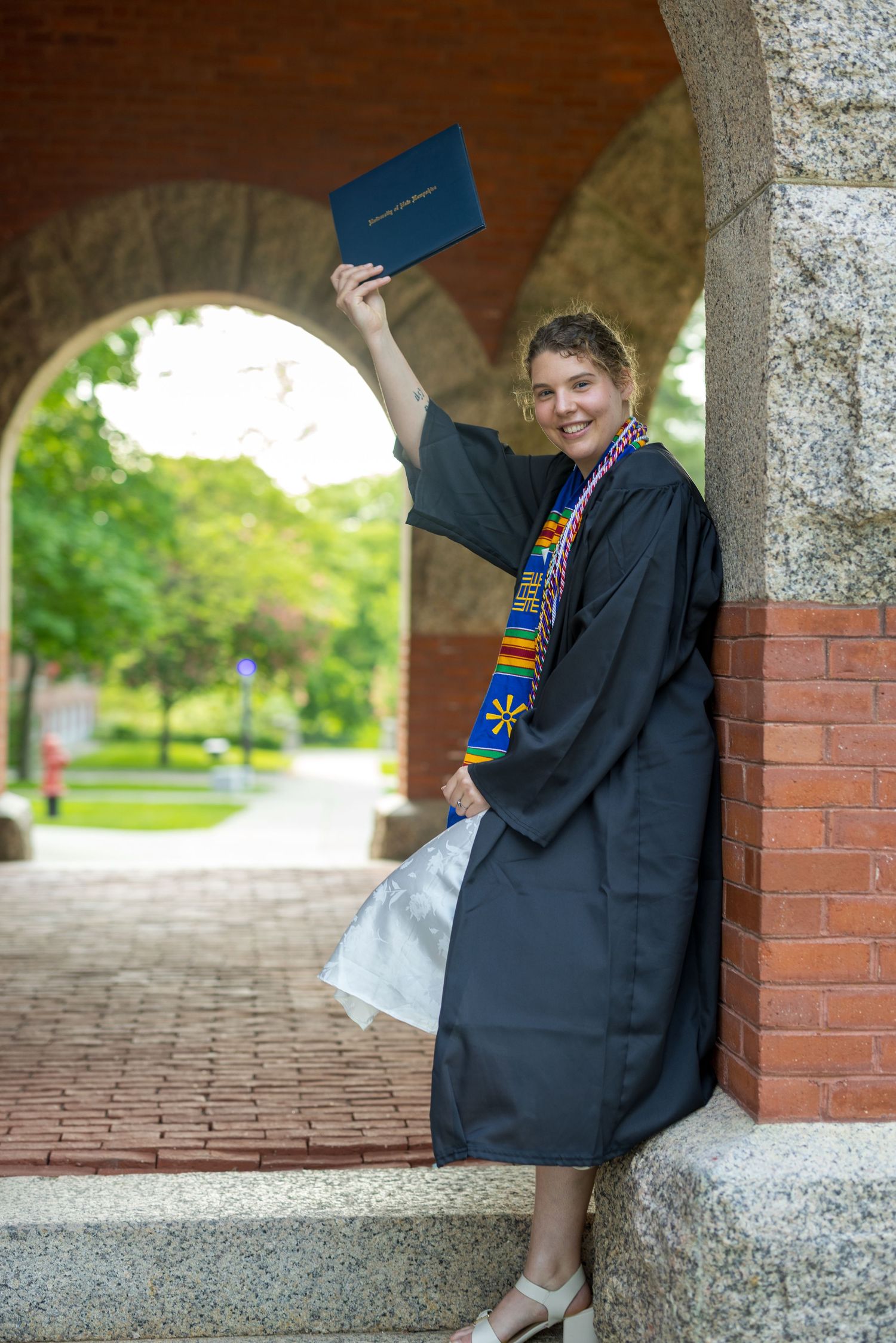 Graduate in black cap and gown holding up diploma while posing under brick archway on campus.