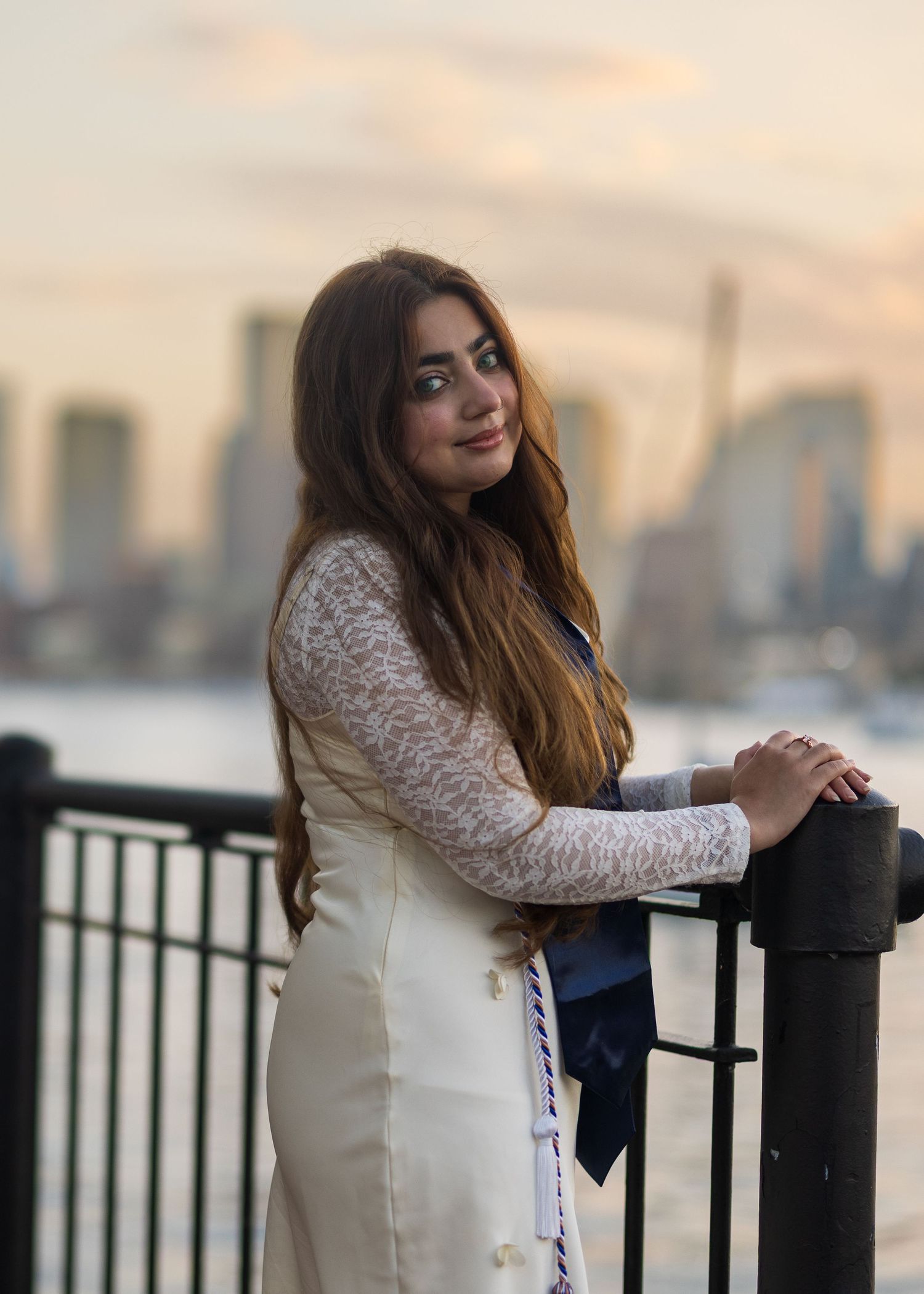 Woman wearing white lace dress leans against waterfront railing at sunset with city skyline in the background.