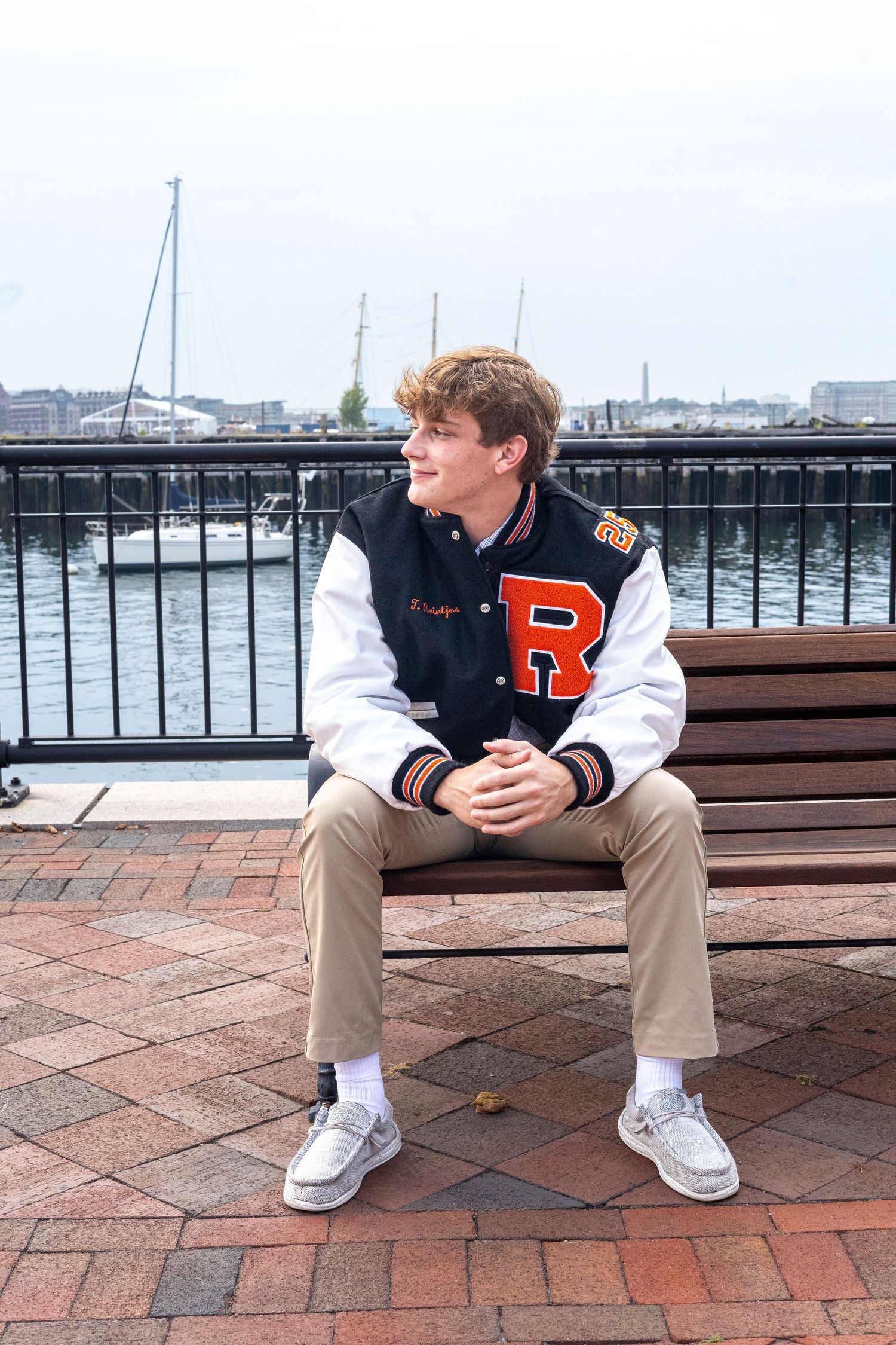 Person in a varsity letterman jacket sits on a bench by a marina with boats and water in the background.