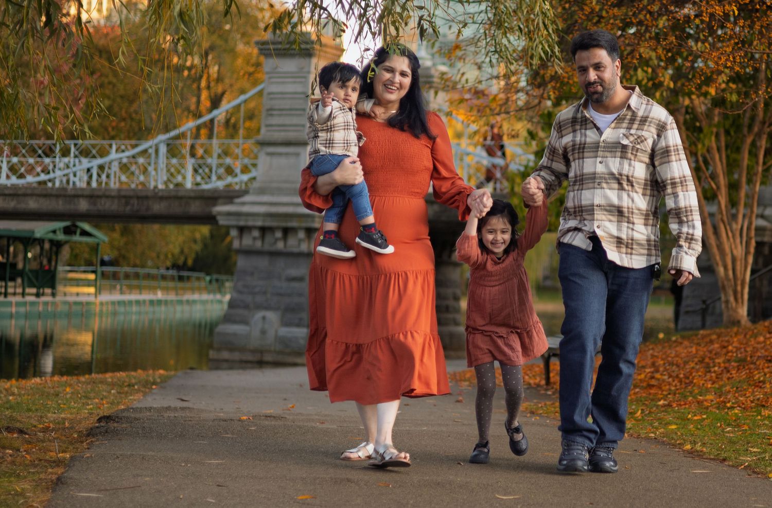 A family walking together on an autumn path by a lake with fall foliage and a bridge in the background.