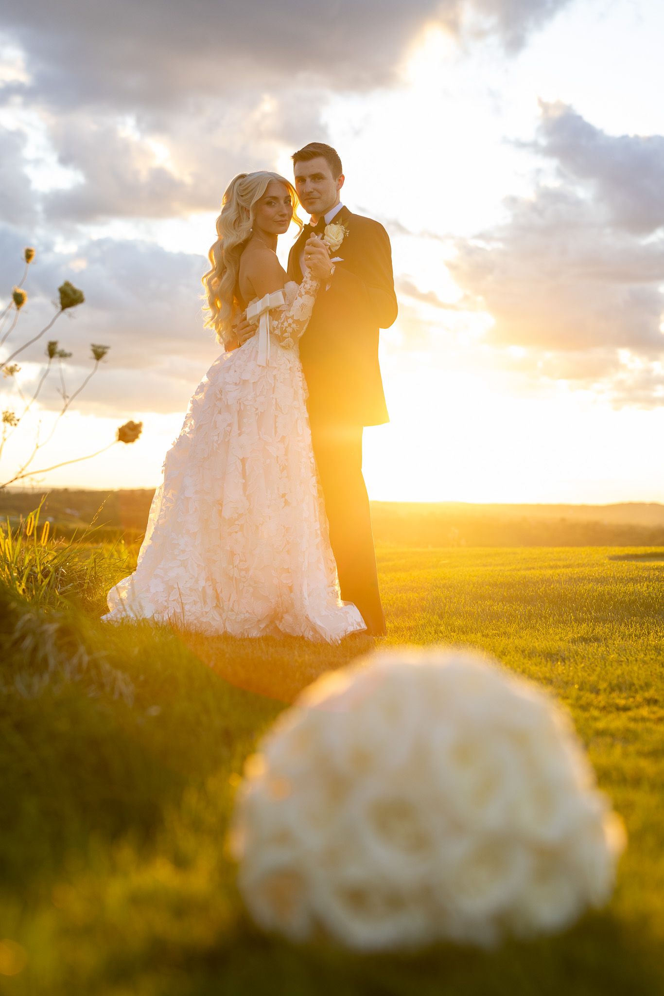Bride and groom embrace at golden sunset on green hill, with white rose bouquet in foreground.