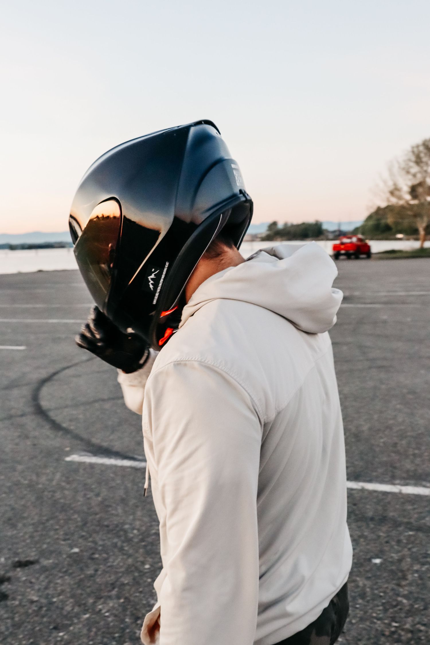 Person wearing motorcycle helmet and white hoodie standing in parking lot with red car in background.
