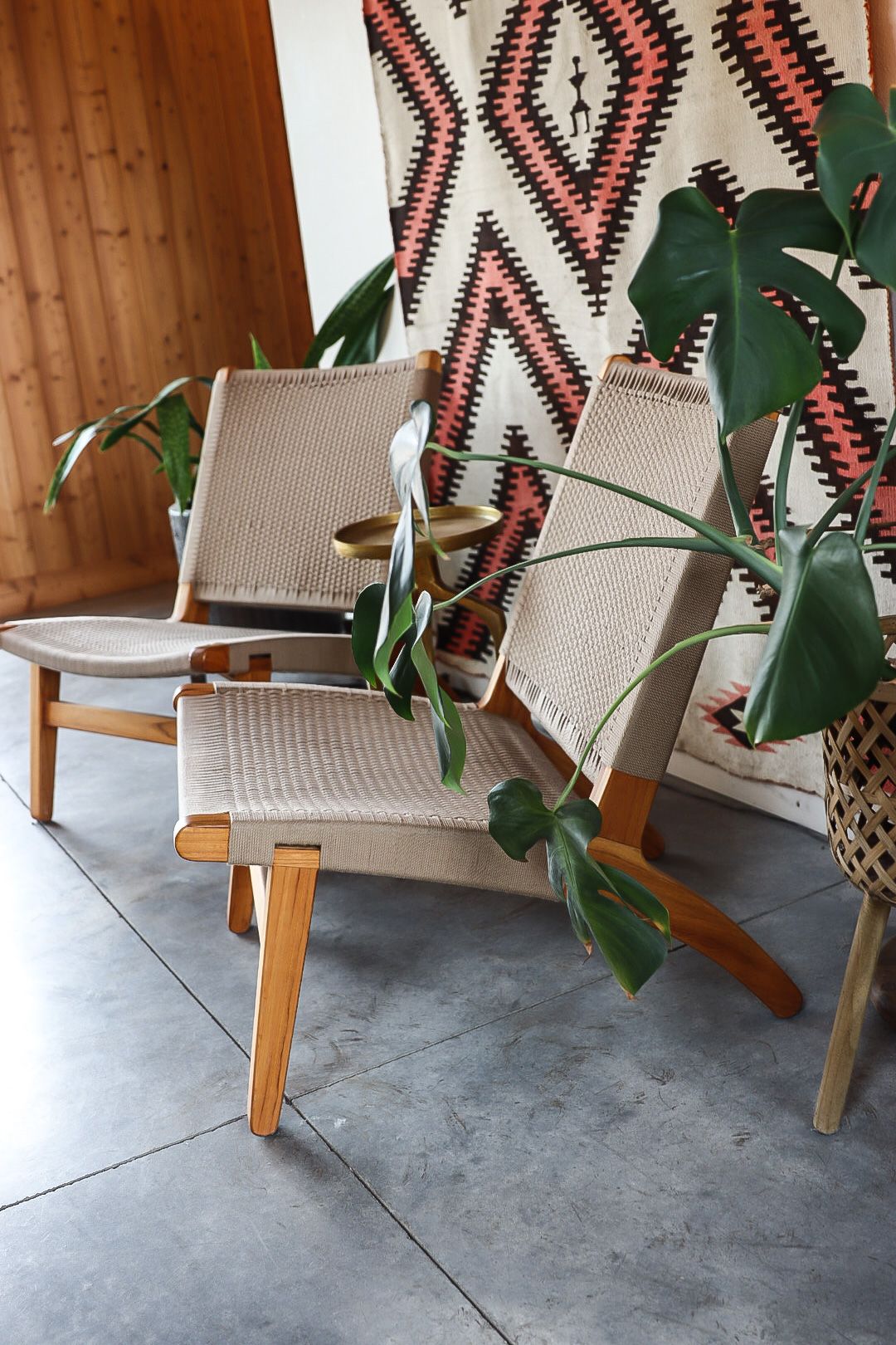 Mid-century modern lounge chairs with woven seats positioned against a geometric wall tapestry with monstera plants.