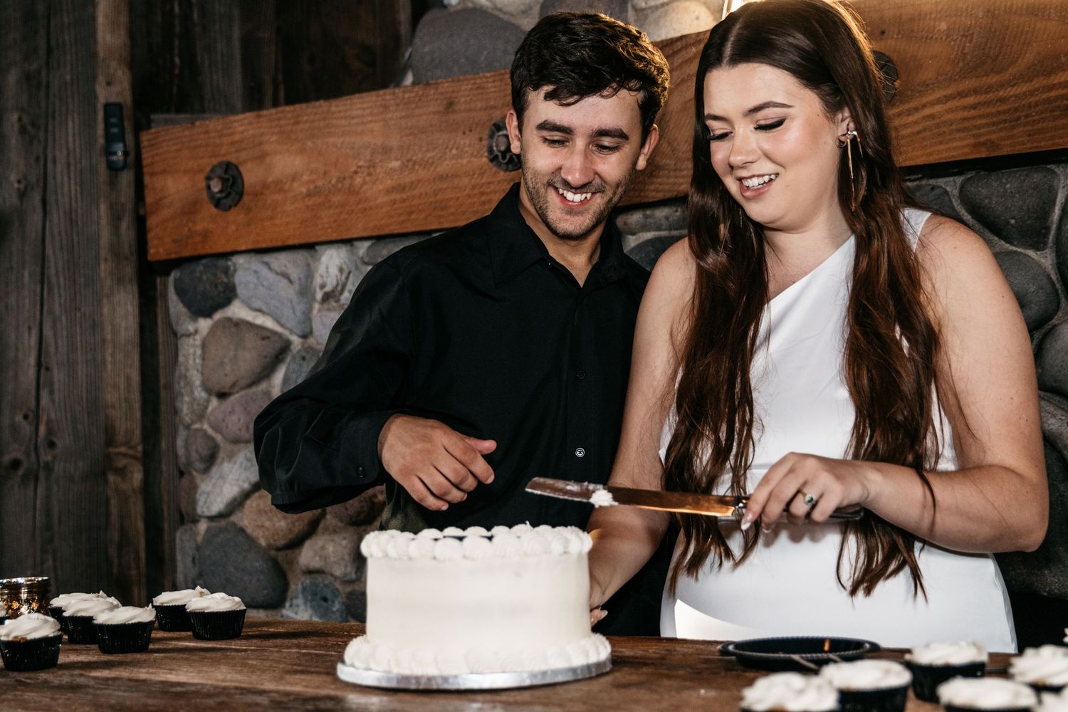 A couple shares a moment while cutting into a white frosted cake together.