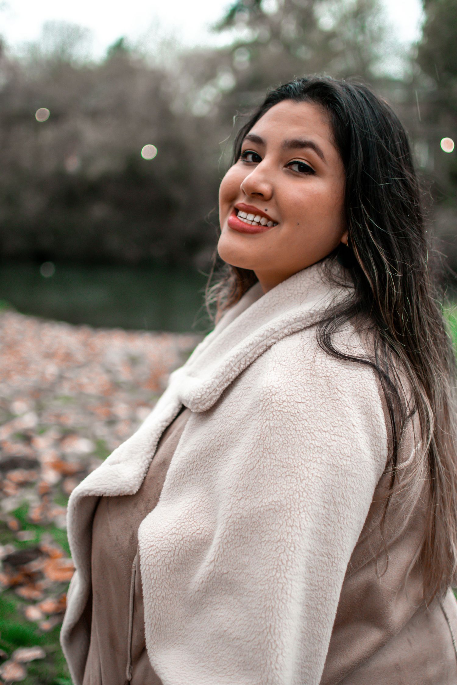 Person in cozy beige teddy coat smiling outdoors during autumn with fallen leaves in background.