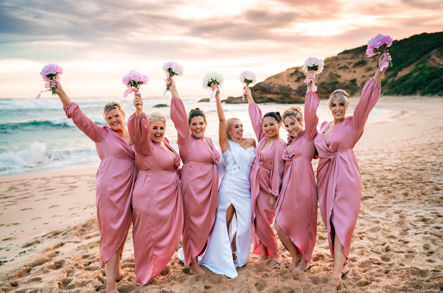 Pink toned bridal party on the beach all with high hair buns with soft curled detail. Bride has long blonde hair in a hollywood waves pony