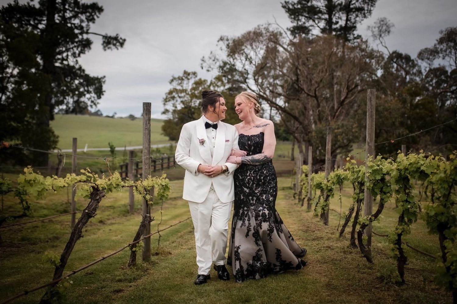 Brides in a beautiful winery field, one with a braided textured high bun and the other with a softer lower bun wearing a black dress