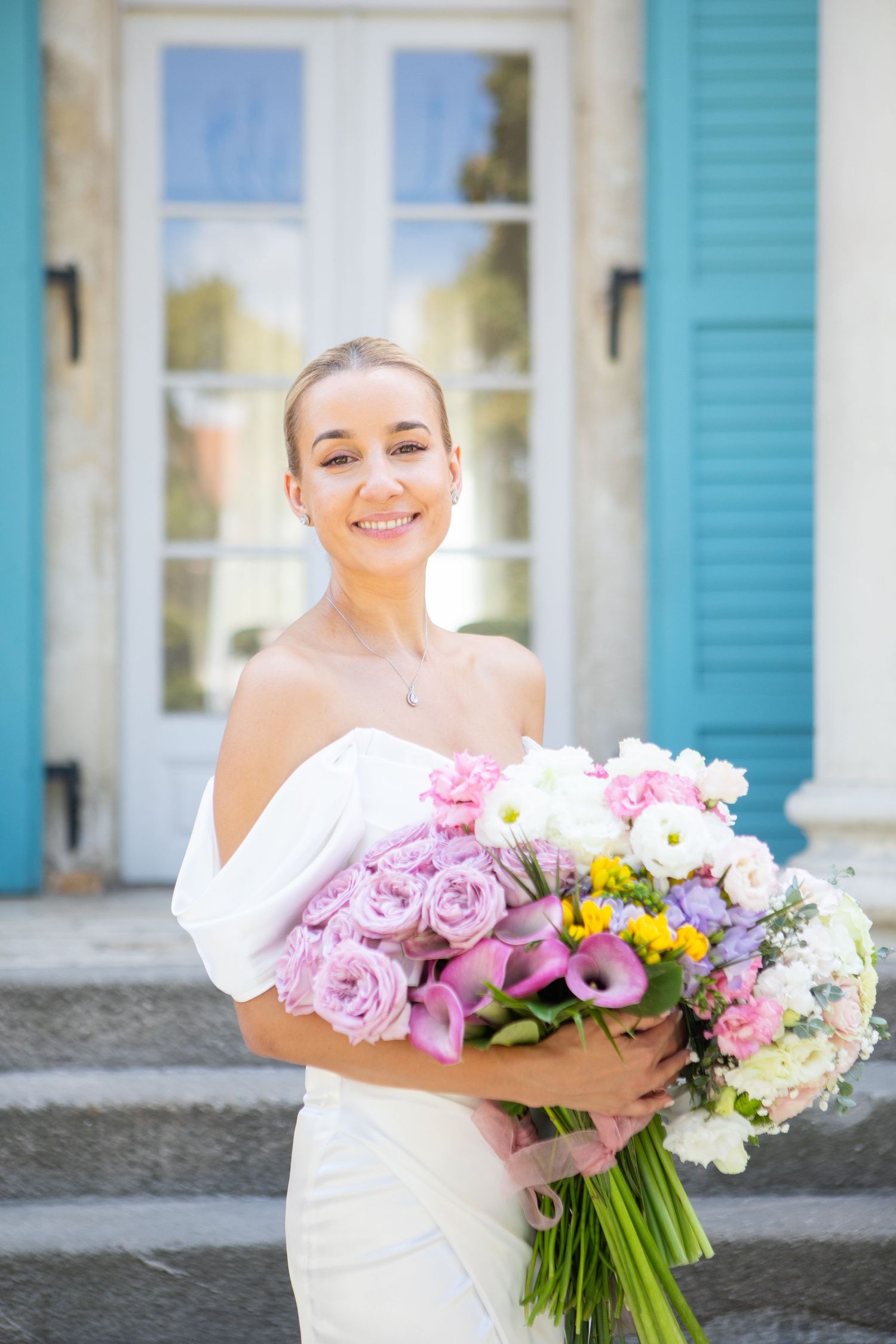 Bride holding a vibrant bouquet of pink and white flowers against turquoise shutters.