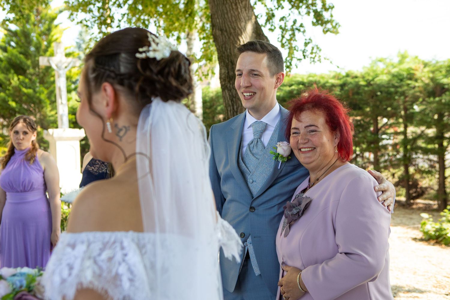 Wedding party members in purple dresses and blue suits gather outdoors for celebratory photos.