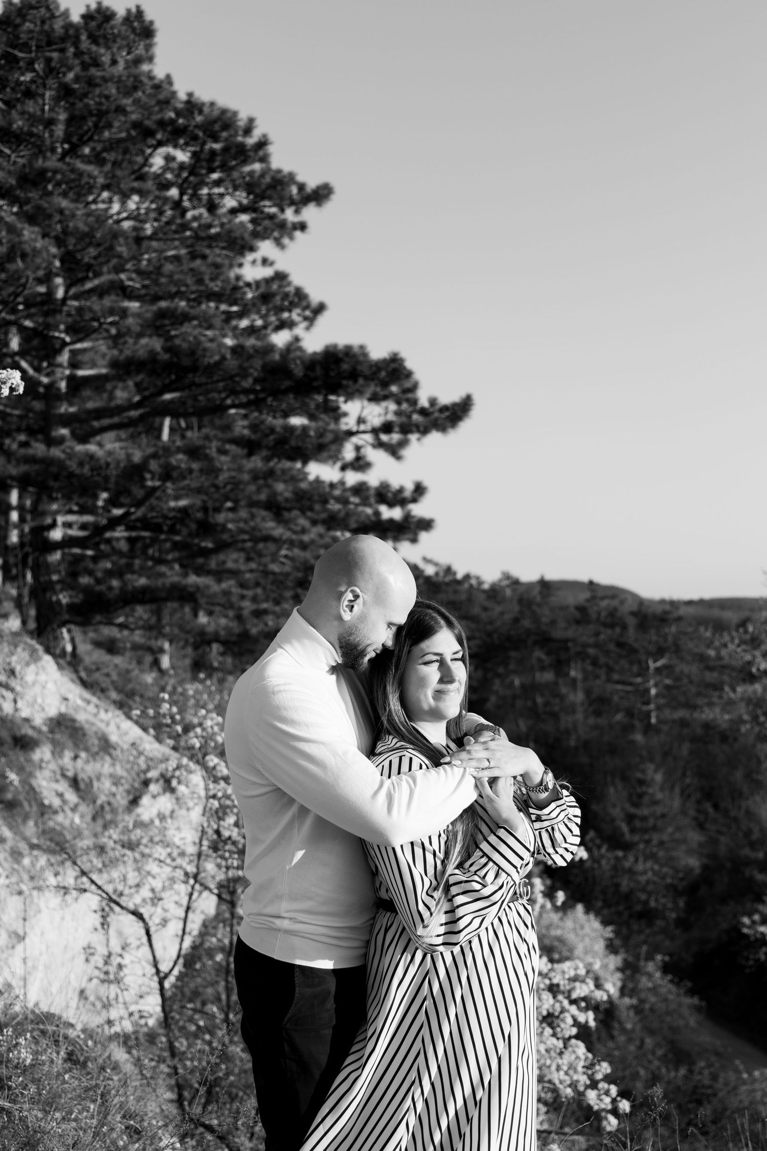 Black and white photo of a couple embracing on a rocky overlook with trees and hills in the background.