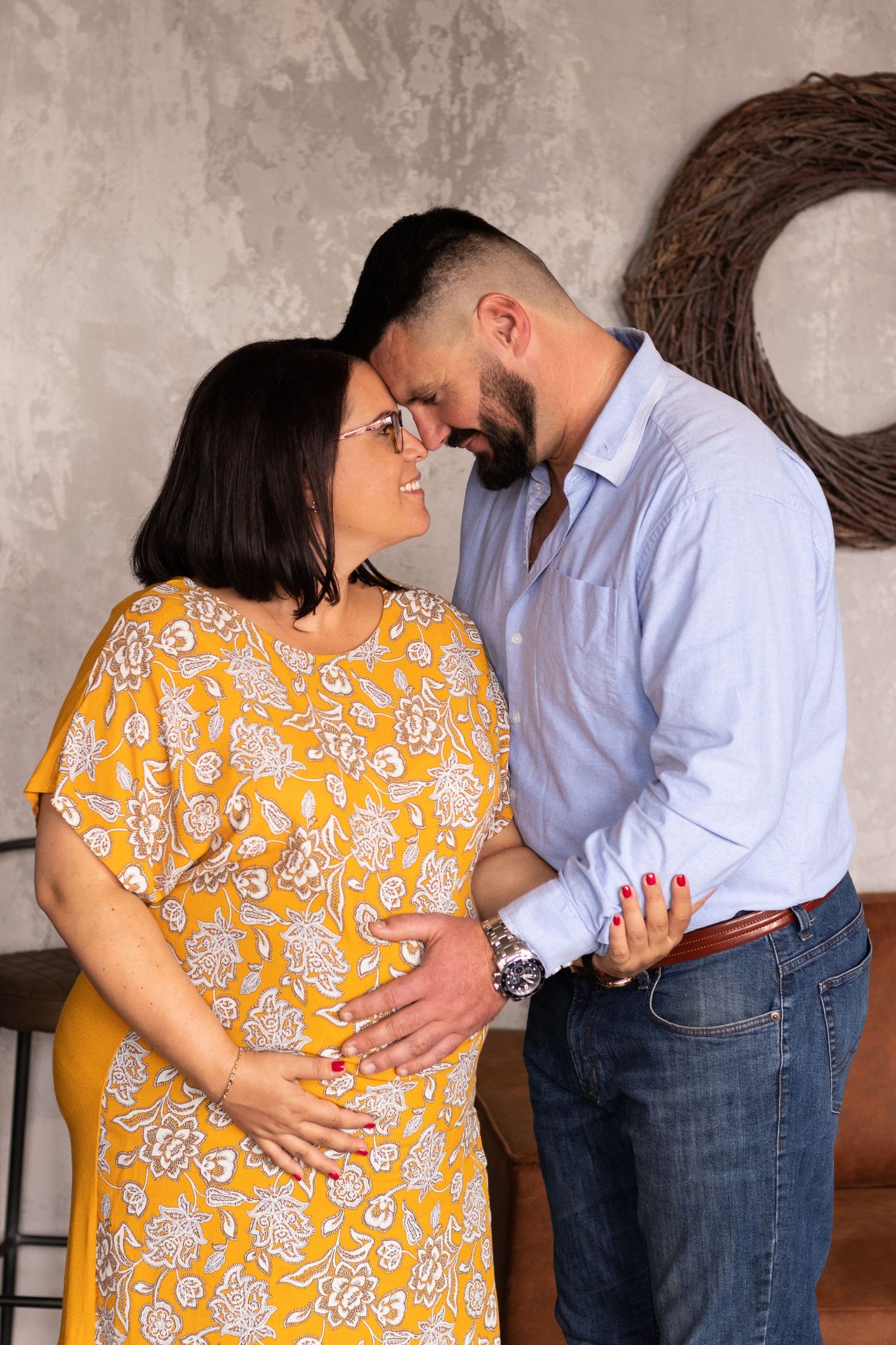 Intimate maternity photos of couple in yellow floral dress and blue shirt against industrial backdrop.