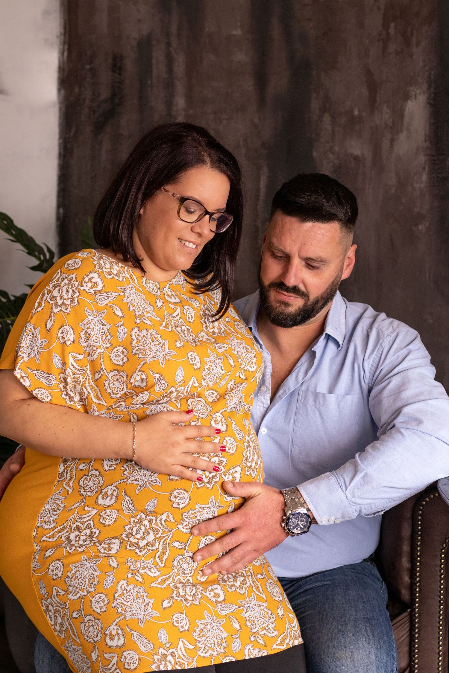Couple in yellow floral dress and blue shirt embrace during maternity photoshoot.