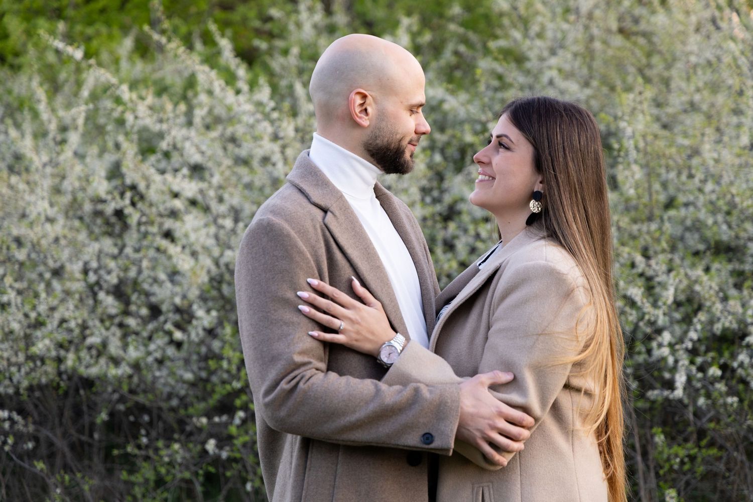 Couple embracing outdoors near blooming white trees, smiling at each other in matching beige coats.