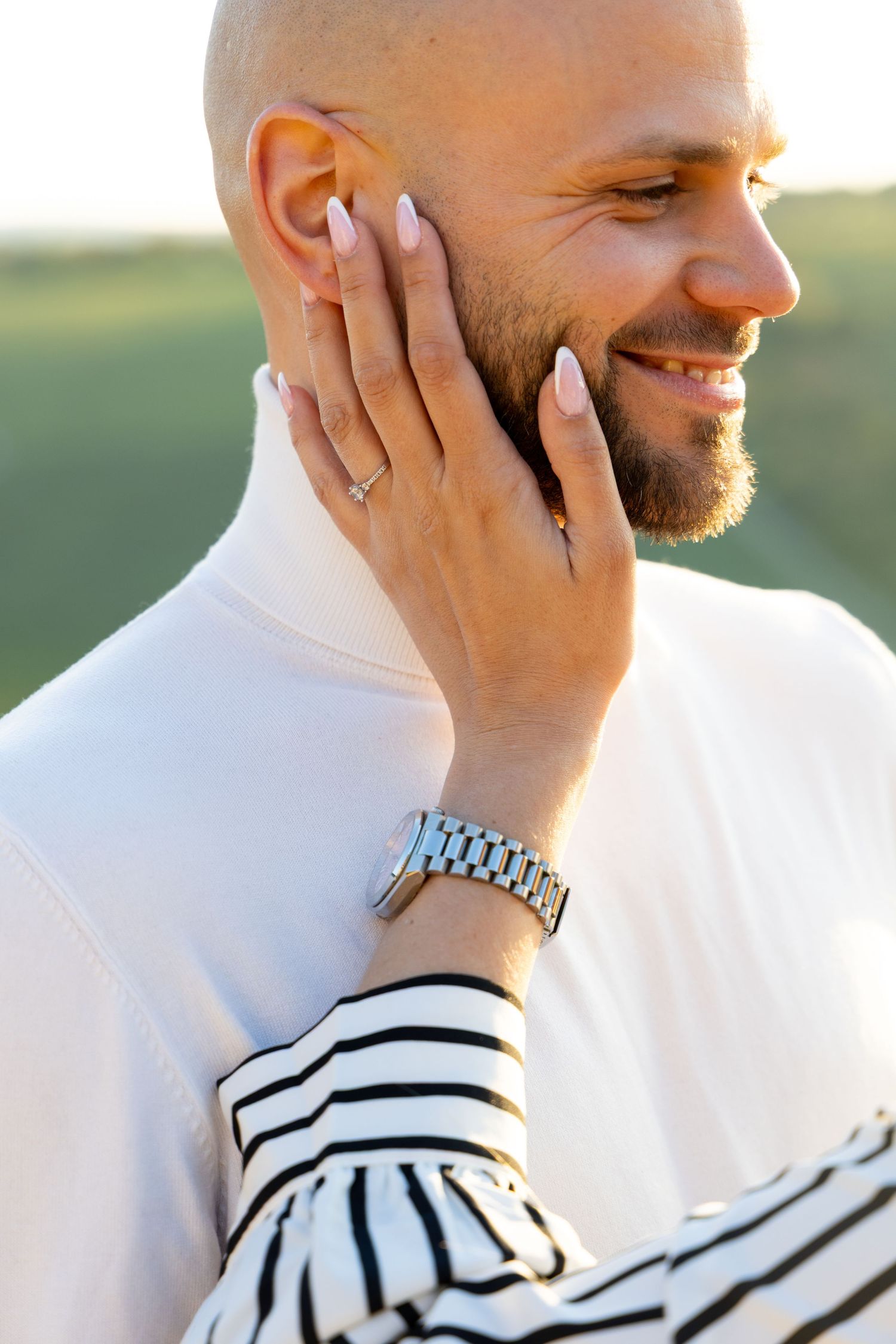 A smiling bald man with a beard gently touched by a hand wearing a silver watch and ring outdoors.