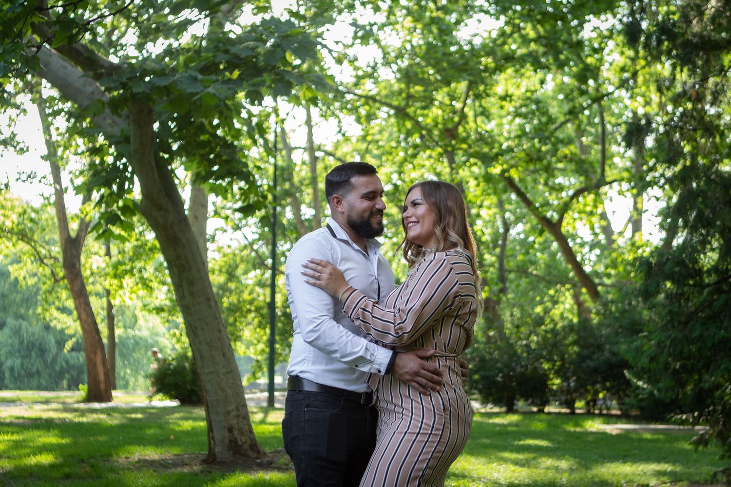 Couple sharing tender moments in a sunlit park with green trees in background.