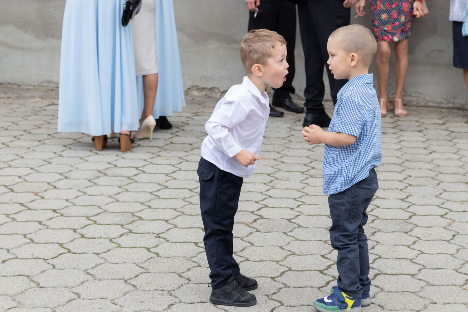 Two young boys in formal attire share an animated conversation on stone pavement.