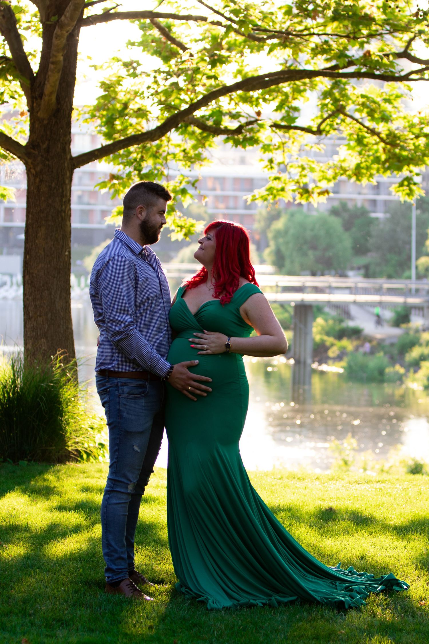 Couple in green dress and blue shirt embrace during sunset maternity photo shoot by river.