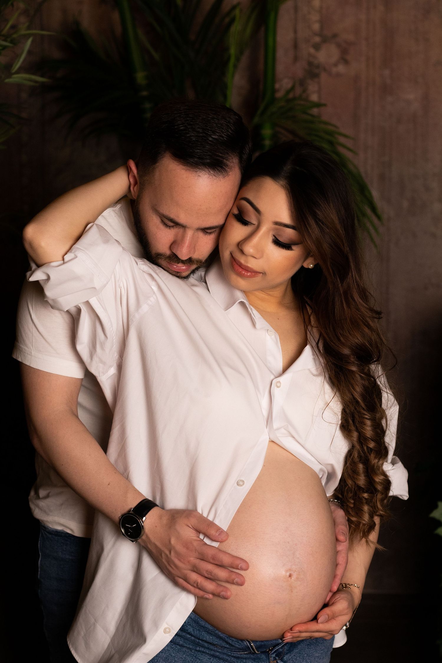 Couple sharing tender moment during pregnancy photoshoot against rustic backdrop.