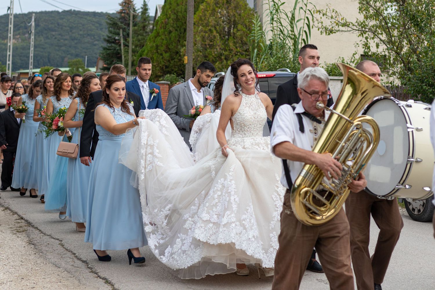 Wedding procession with brass band musician leading bridesmaids in blue dresses down street.