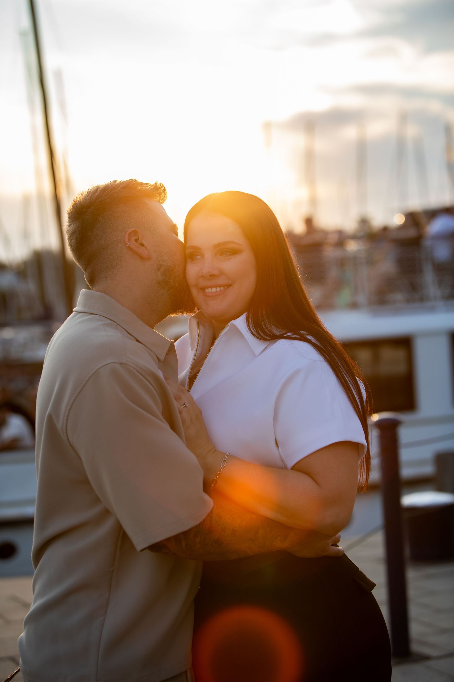 Couple sharing a romantic sunset kiss at a marina, woman in white top smiling warmly with golden backlight.