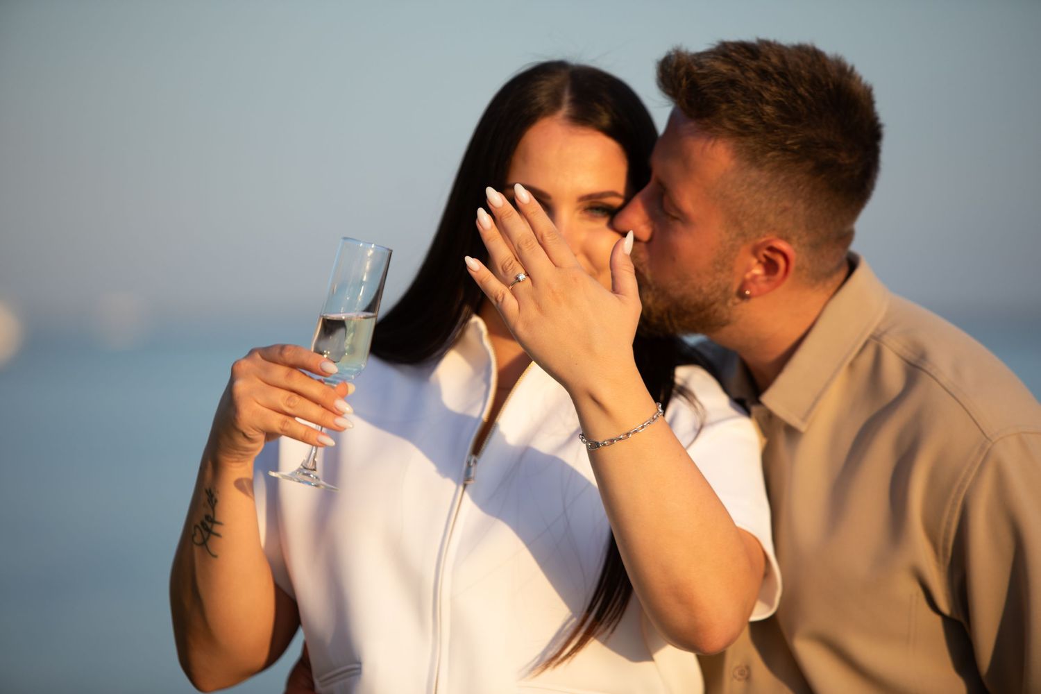 Couple sharing a romantic kiss outdoors at sunset, woman holding champagne glass with engagement ring visible.