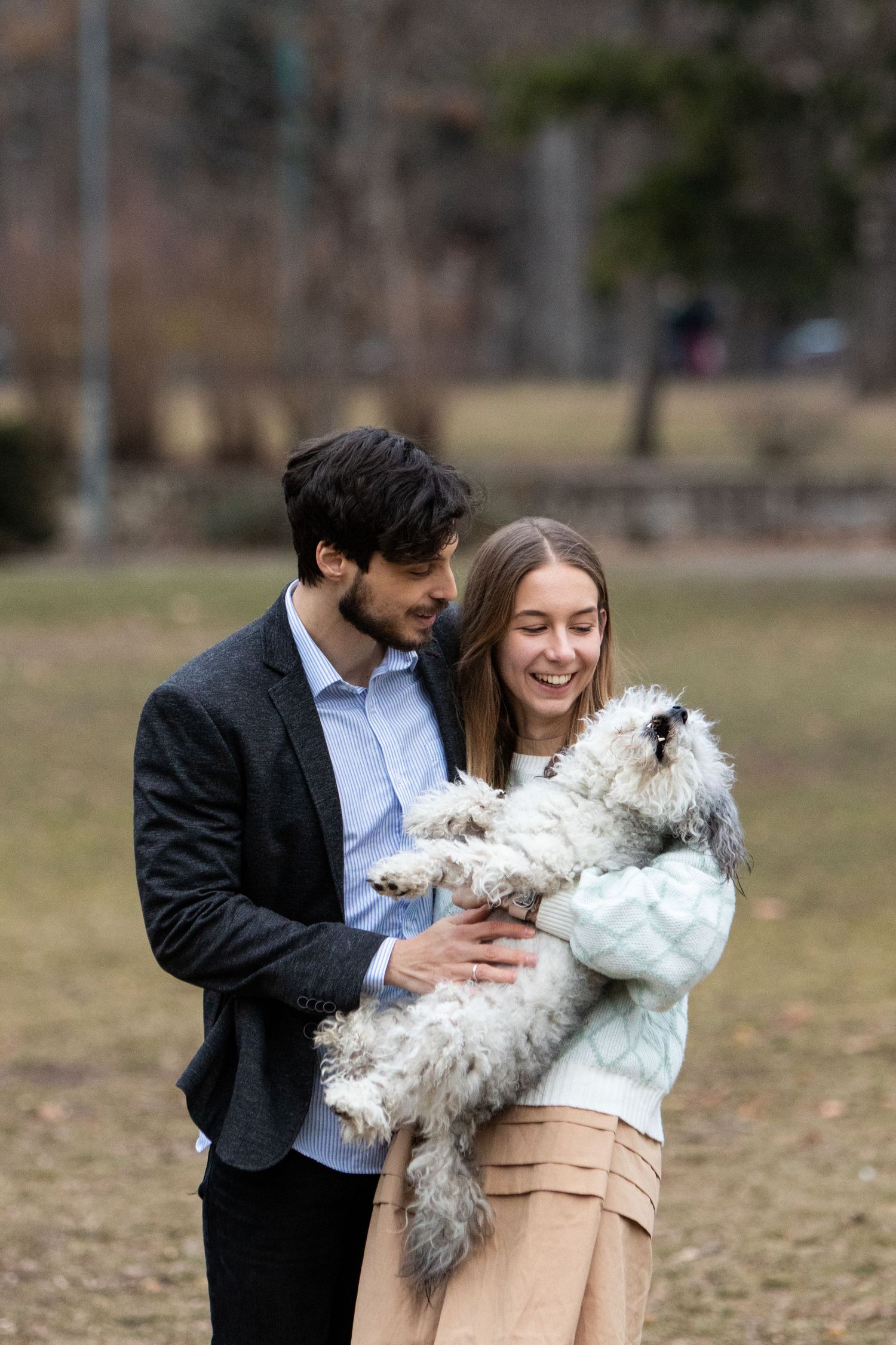 A couple plays with their white fluffy dog in a park setting.