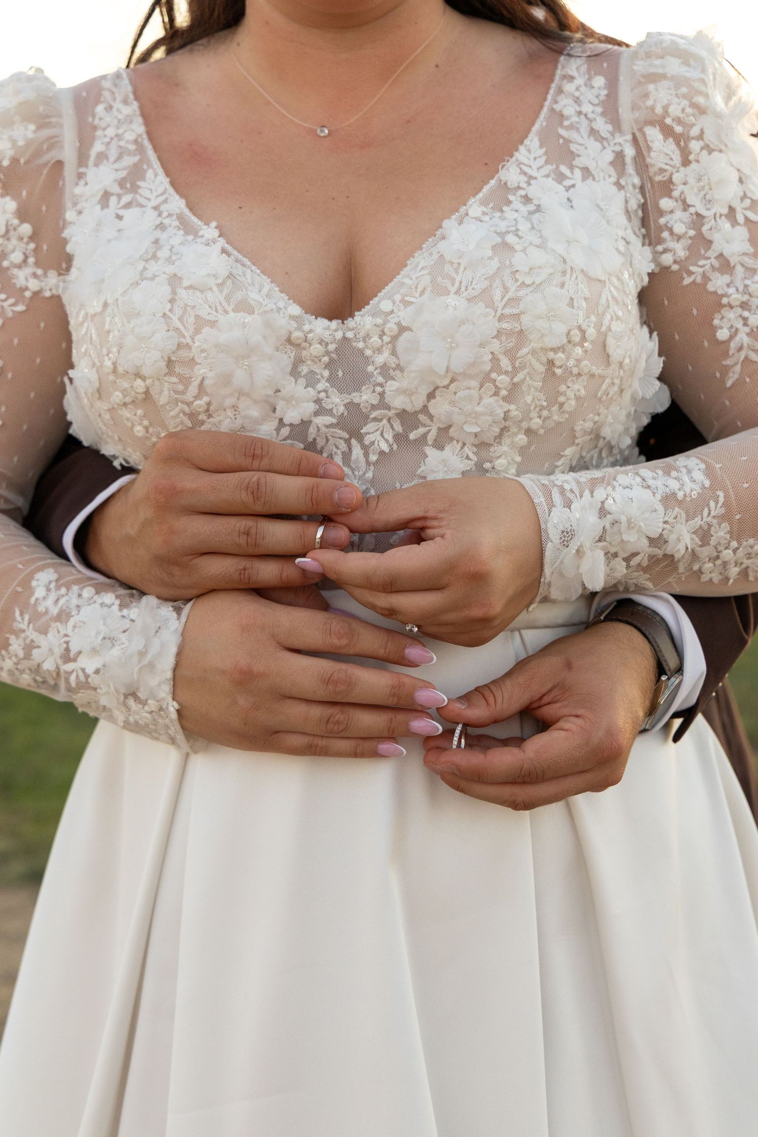 Close-up detail of lace wedding dress bodice and hands with rings.