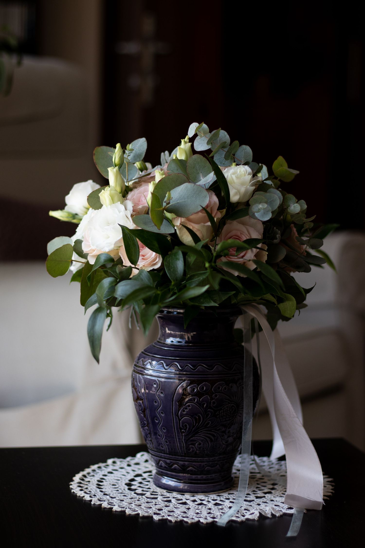 Elegant floral arrangement in a dark decorative vase sits atop a white lace doily on a black surface.