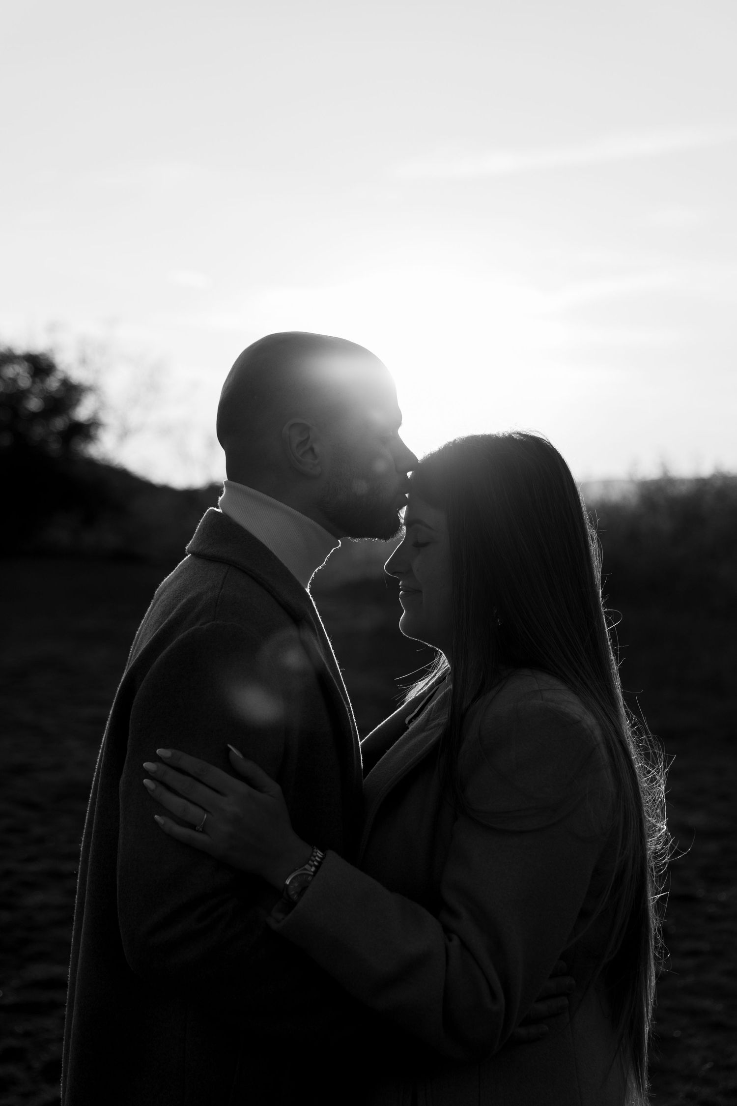 Black and white romantic couple portrait backlit by sunset, embracing closely outdoors.