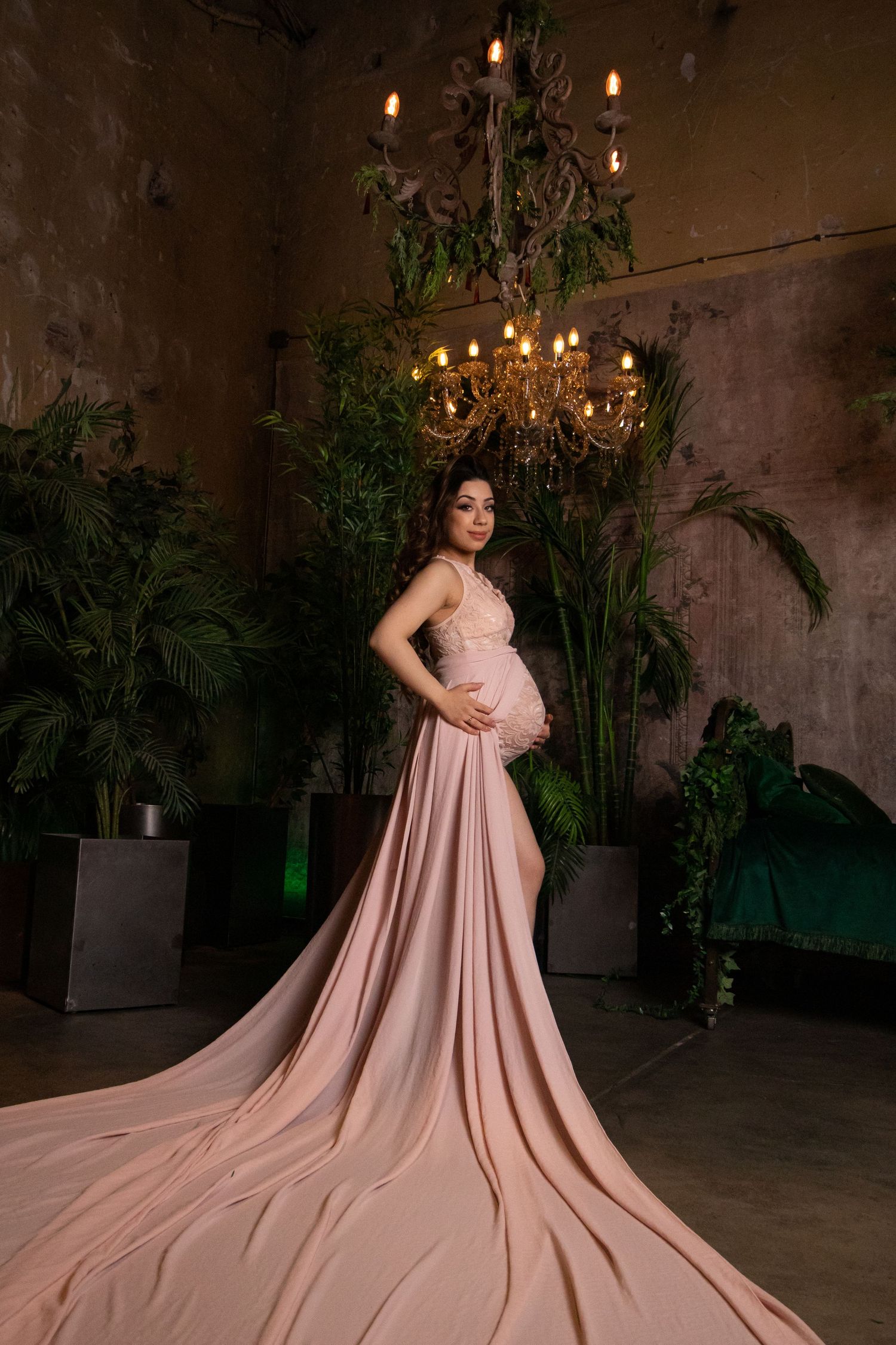 Maternity photoshoot featuring flowing pink gown in dramatic indoor setting with greenery.