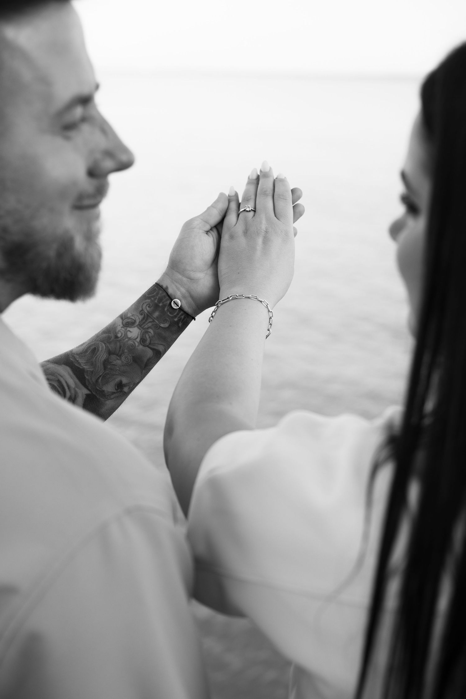 Black and white photo of a couple holding hands, showing an engagement ring, with a tattooed man smiling near water.