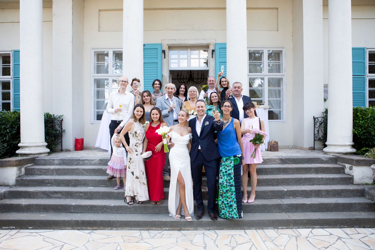 Large wedding party gathered on white steps of an elegant building with turquoise shutters.