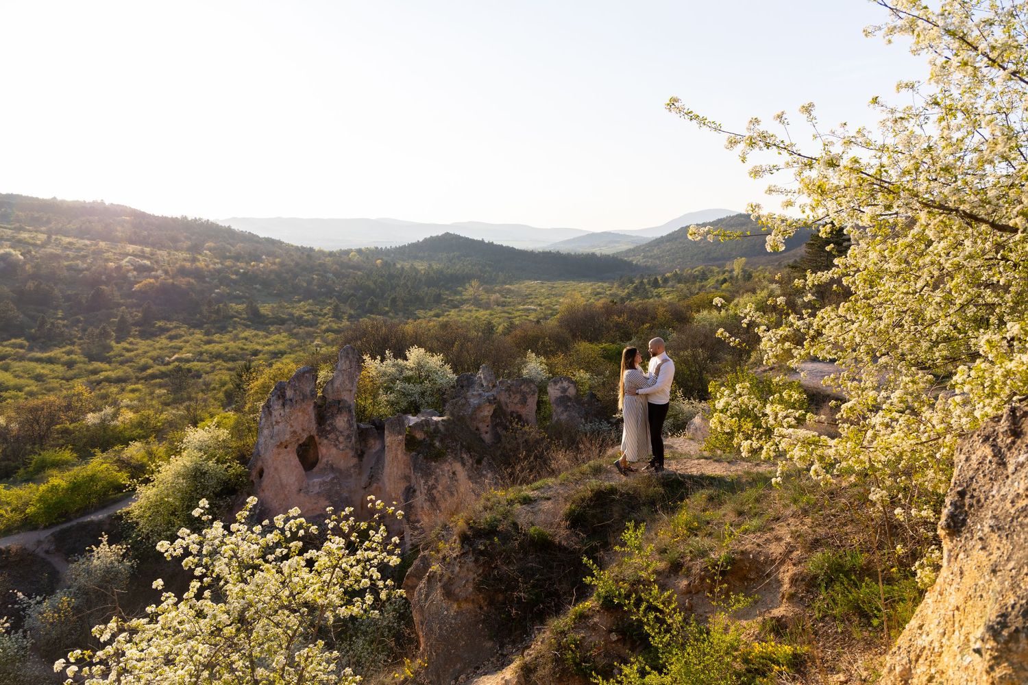 Couple standing on rocky cliff edge surrounded by blooming spring trees and lush green valley landscape at sunset.