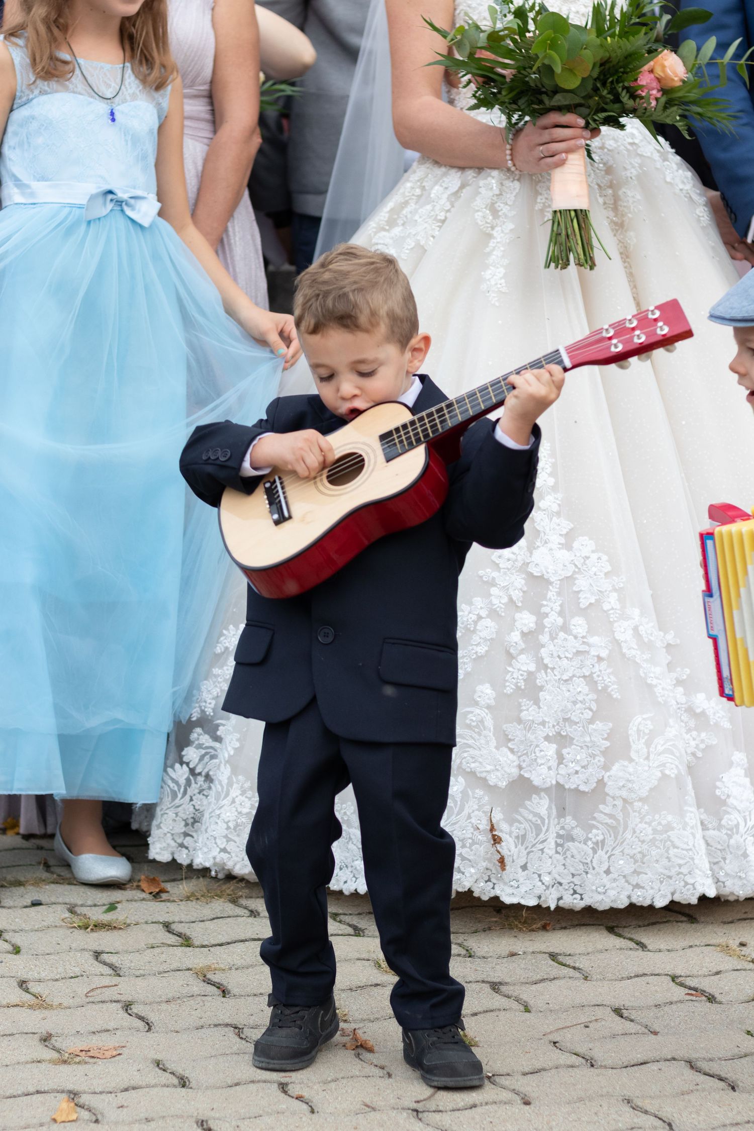 Young boy in formal suit plays small guitar next to wedding dress and light blue bridesmaid dress.
