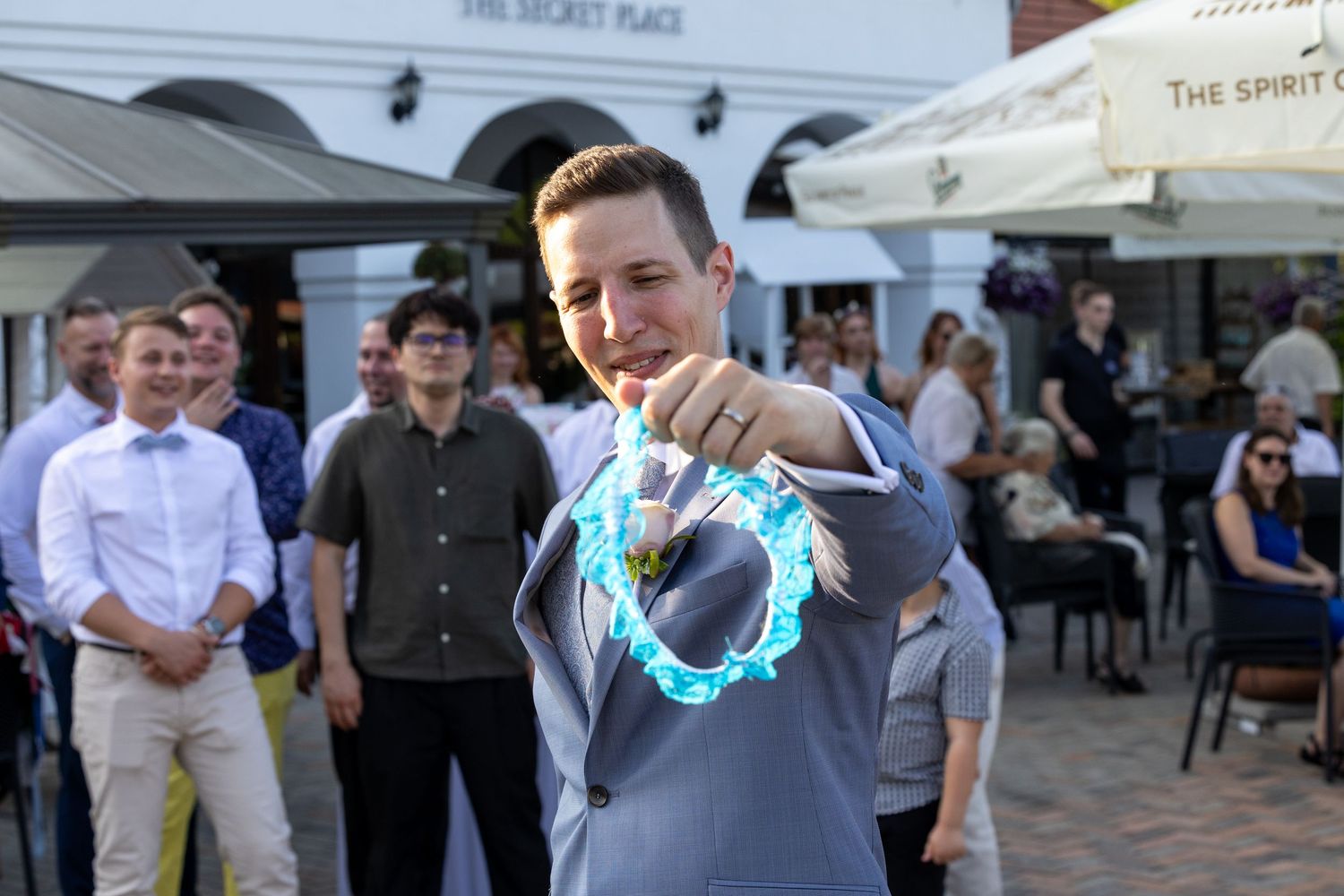 Groom in grey suit holds glowing blue ring at outdoor reception.