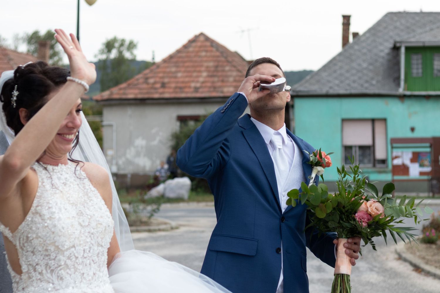 Newlyweds share laughter while holding bouquets outdoors near colorful buildings.
