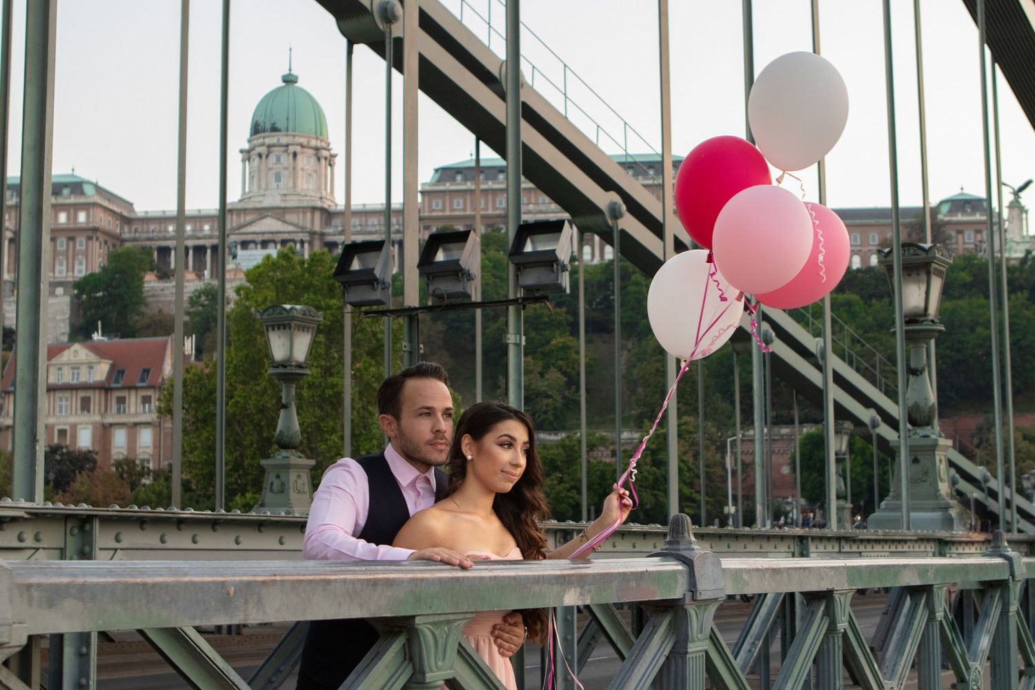 Couple holding pink and white balloons on a bridge with city architecture in the background.