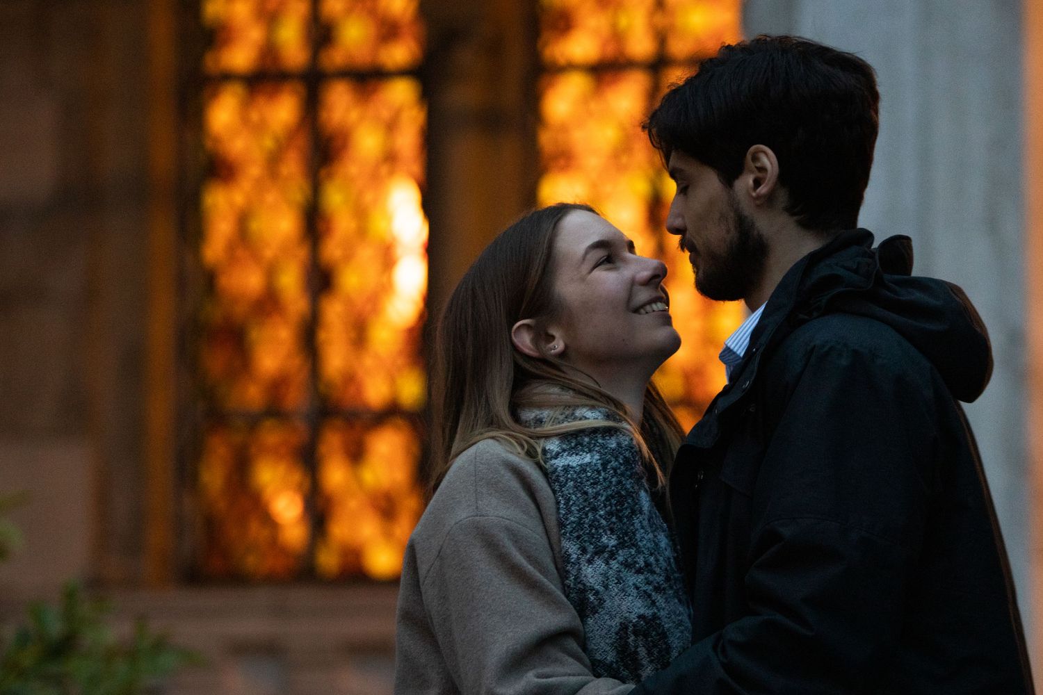 Couple sharing romantic moment in front of stained glass window with warm orange glow.