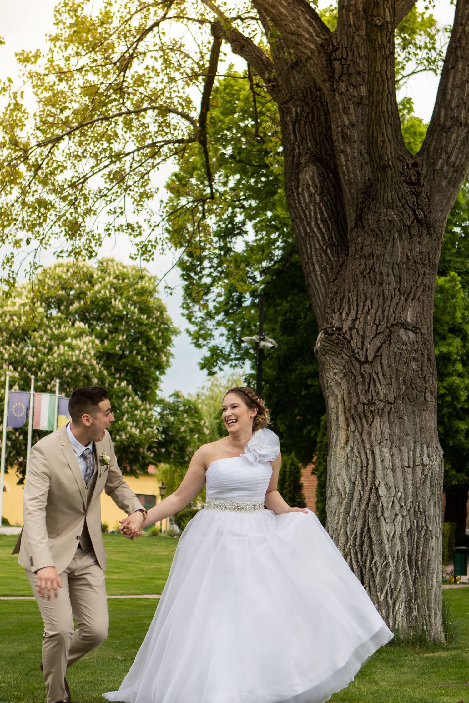 A joyful wedding couple shares a playful moment among tall trees in a park setting.