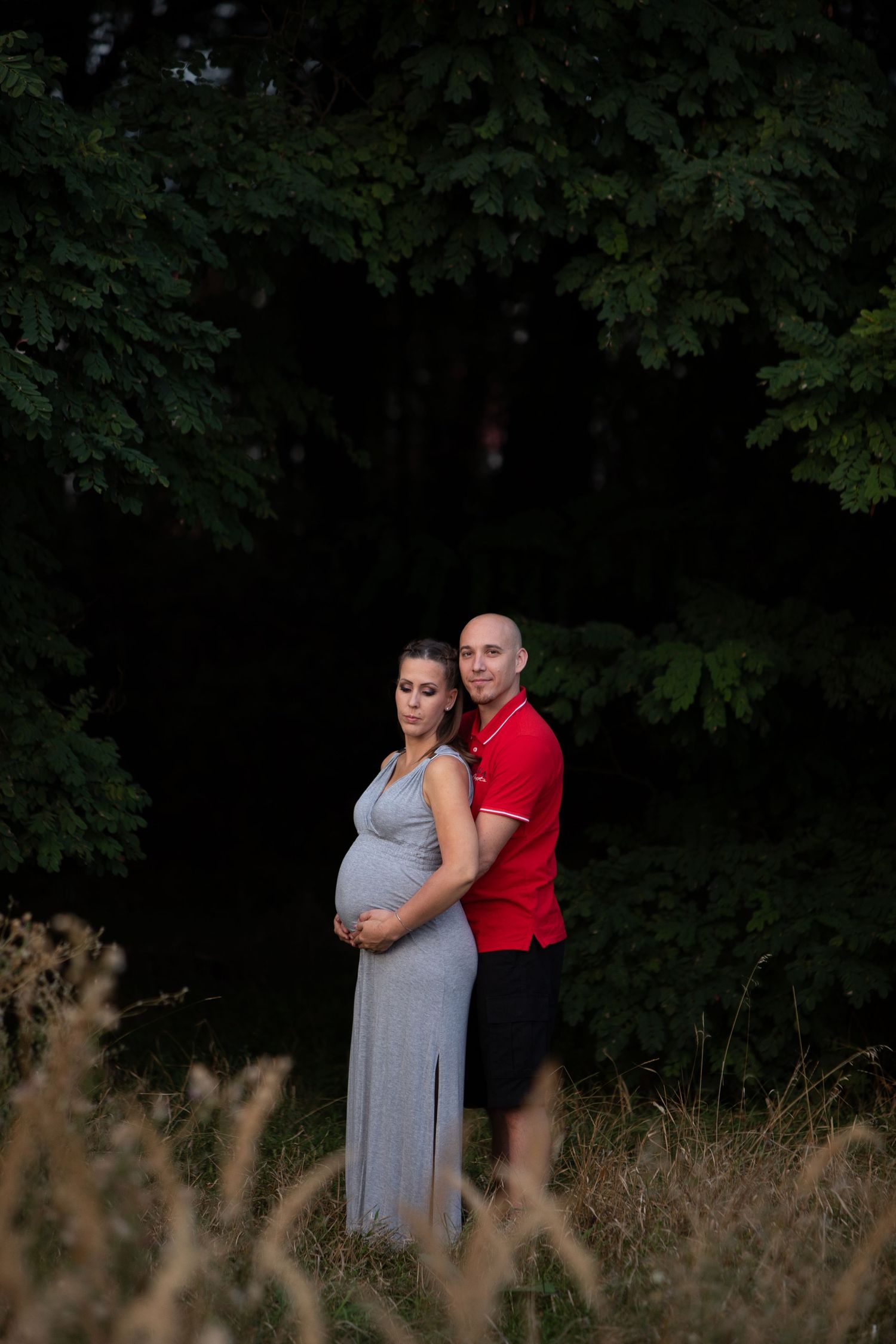 Couple embraces in a romantic outdoor photoshoot with trees and tall grass in the background.