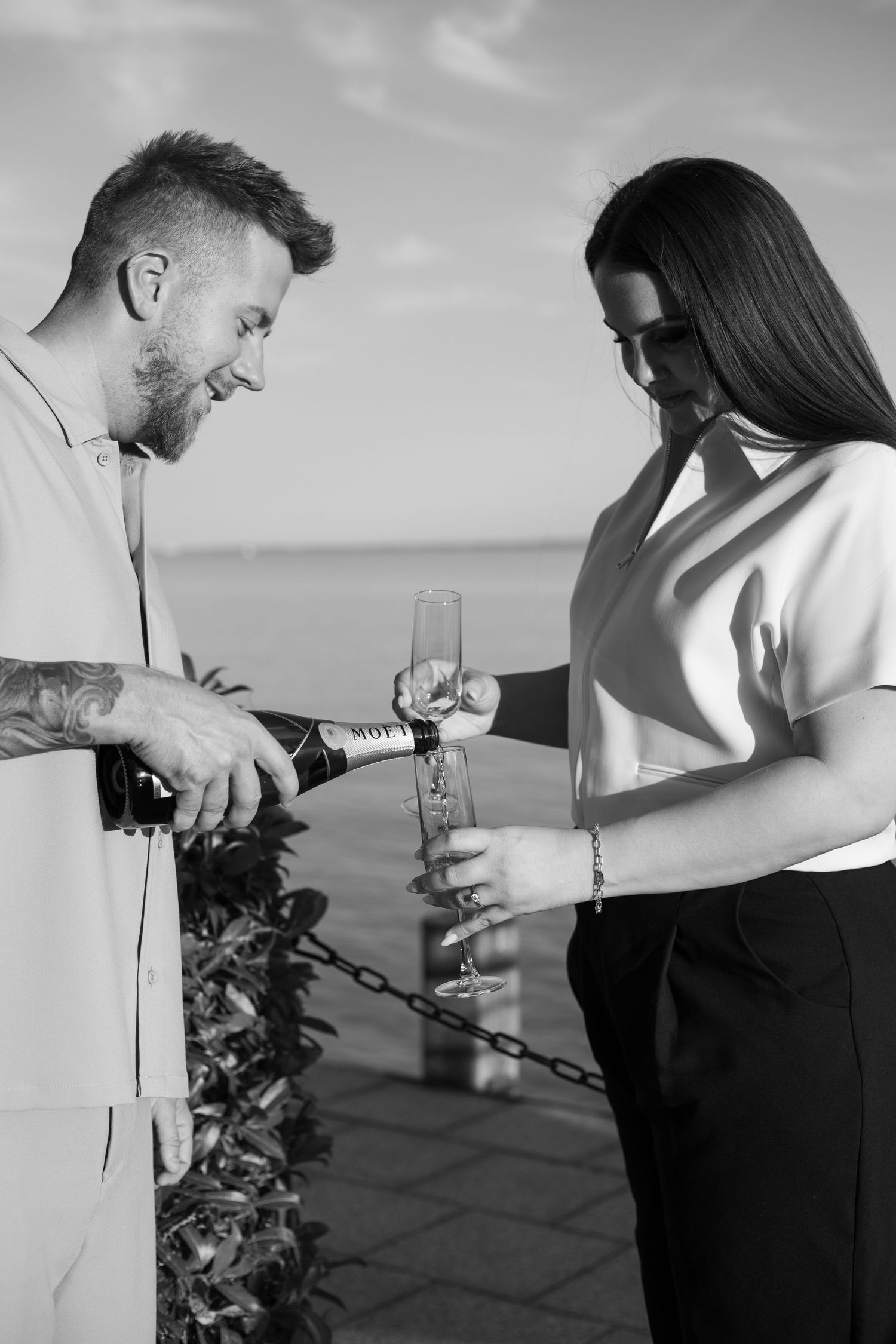 Man pouring champagne into a glass held by a woman outdoors near the sea.